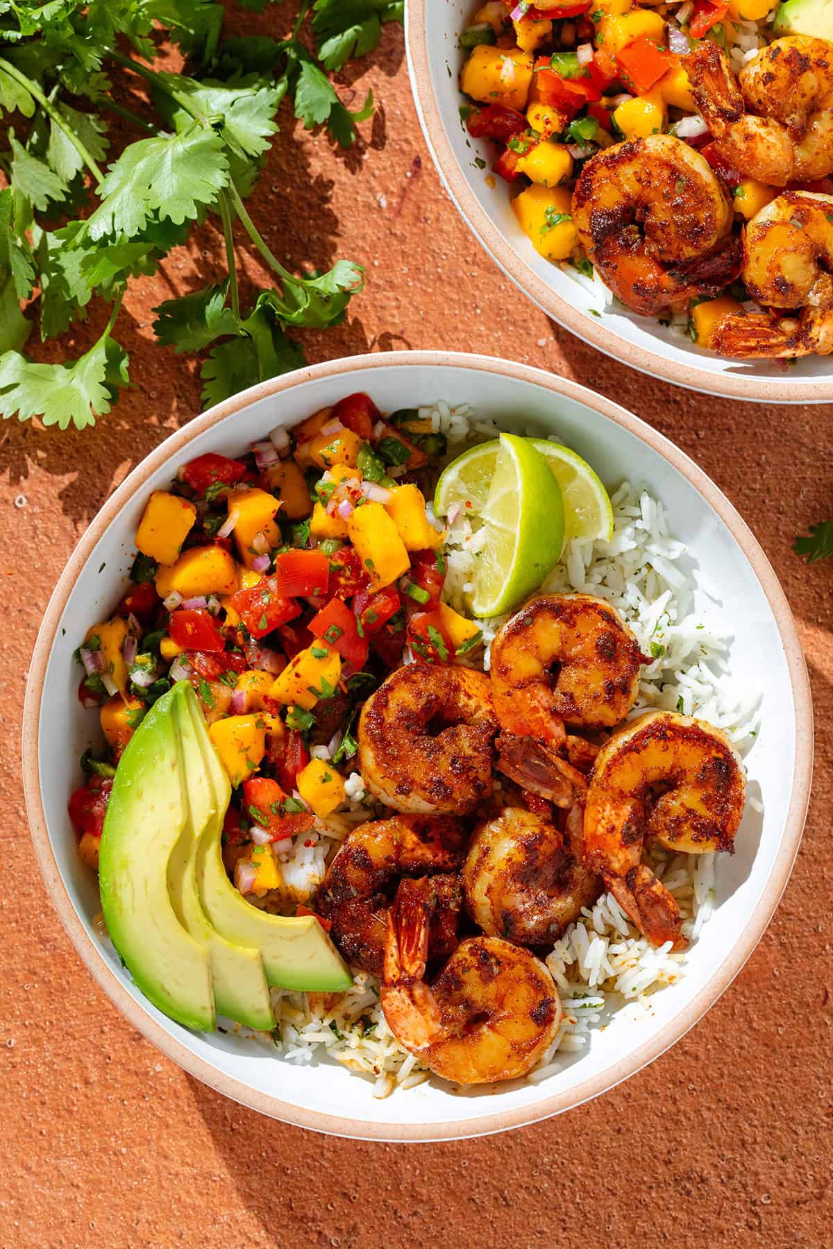 A high protein bowl of rice topped with grilled shrimp, avocado slices, mango salsa, and lime wedges, with fresh cilantro on the side—perfect for a refreshing summer meal.