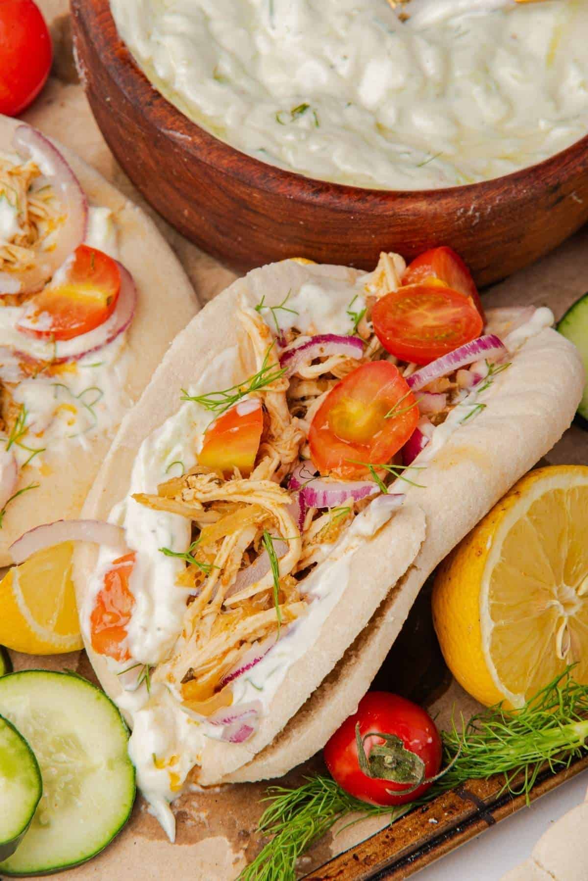 Pita bread filled with slow cooker shredded chicken, cherry tomatoes, red onions, and tzatziki sauce, surrounded by cucumber slices, lemon halves, and a bowl of tzatziki—perfect for easy summer recipes.