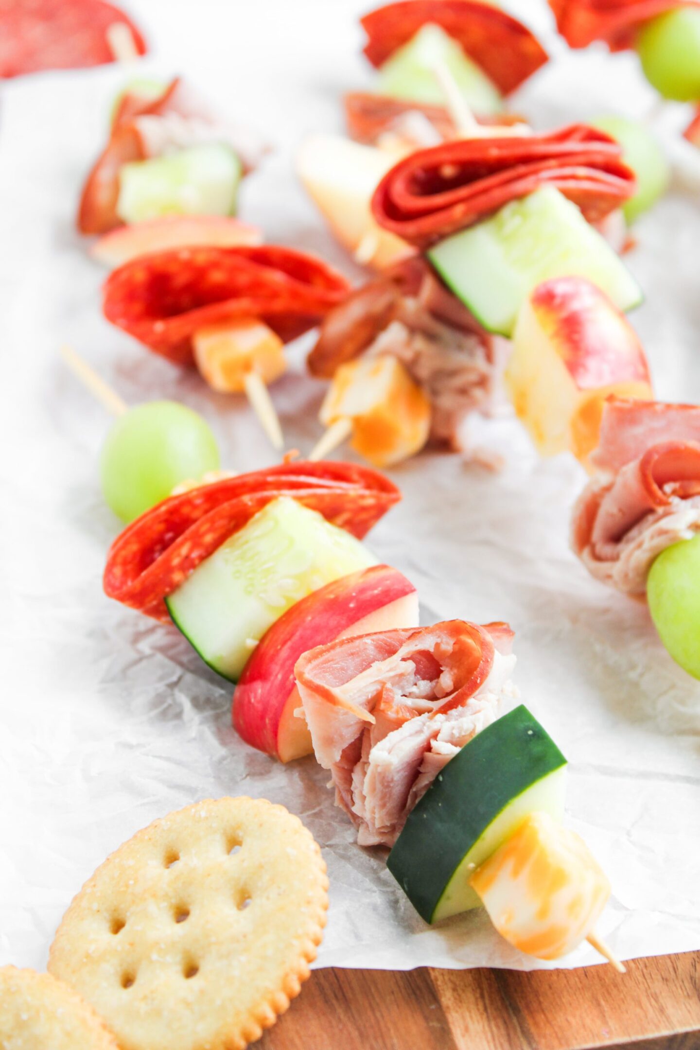 Skewers with sliced meats, cheese cubes, cucumber, apple, and grapes arranged on parchment paper, with round crackers nearby—perfect for cold beach lunch ideas.