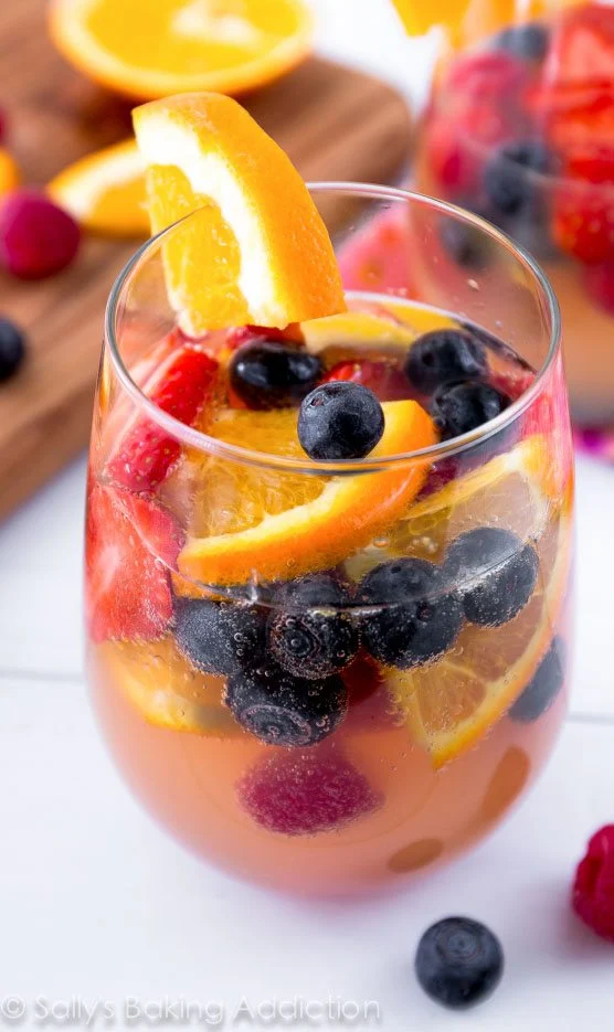 A glass of summer sangria with orange slices, blueberries, strawberries, and raspberries sits in front of more fruit and another drink in the background.