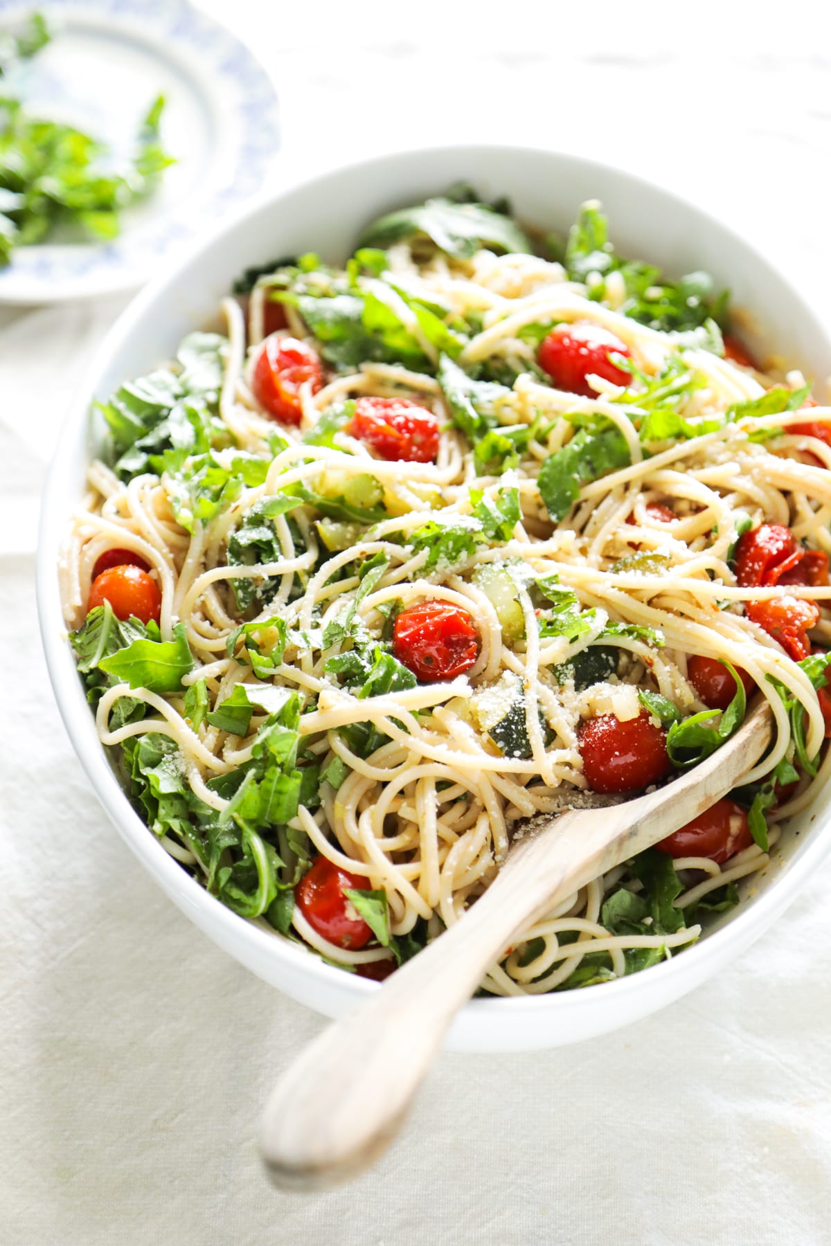 A white bowl of spaghetti with cherry tomatoes, fresh greens, and grated cheese, served with a wooden spoon—perfect for easy pasta recipes and delightful summer pasta recipes.