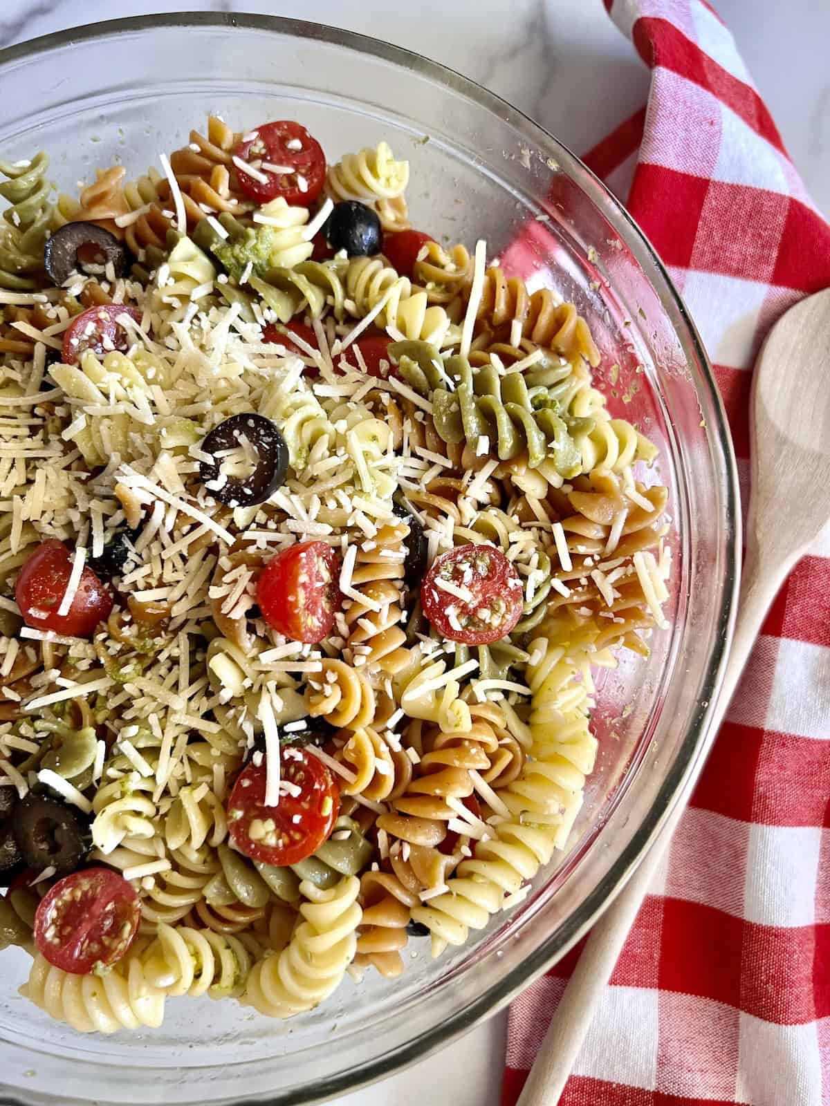 A glass bowl of tri-color rotini pasta salad with cherry tomatoes, black olives, and shredded cheese makes a colorful side dish—perfect for a graduation party—served beside a wooden spoon and a red and white checkered napkin.