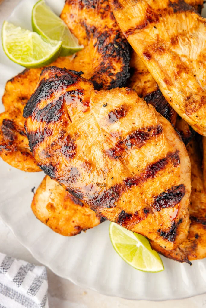 Grilled chicken breasts with char marks are served on a white plate, garnished with lime wedges—perfect for a light summer meal.