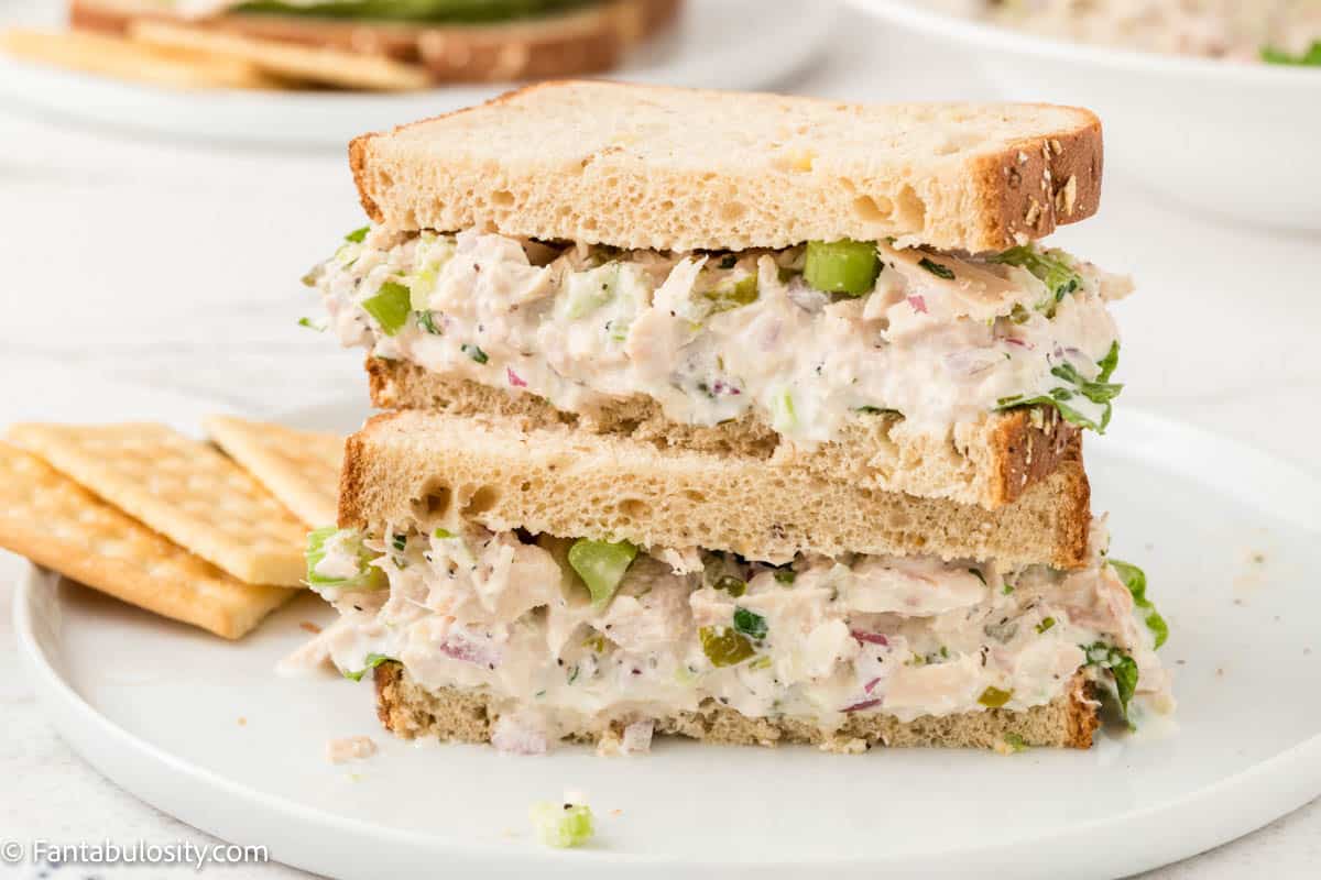 A stacked chicken salad sandwich with lettuce on sliced bread sits on a white plate next to two saltine crackers—perfect for easy cold beach lunch ideas.
