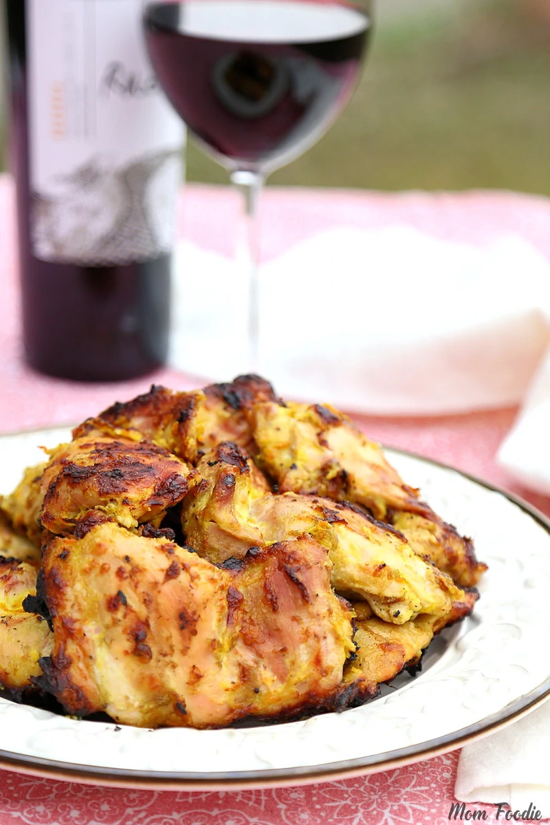 A plate of grilled chicken, perfect for summer, sits on a white plate with a glass of red wine and a wine bottle in the background.