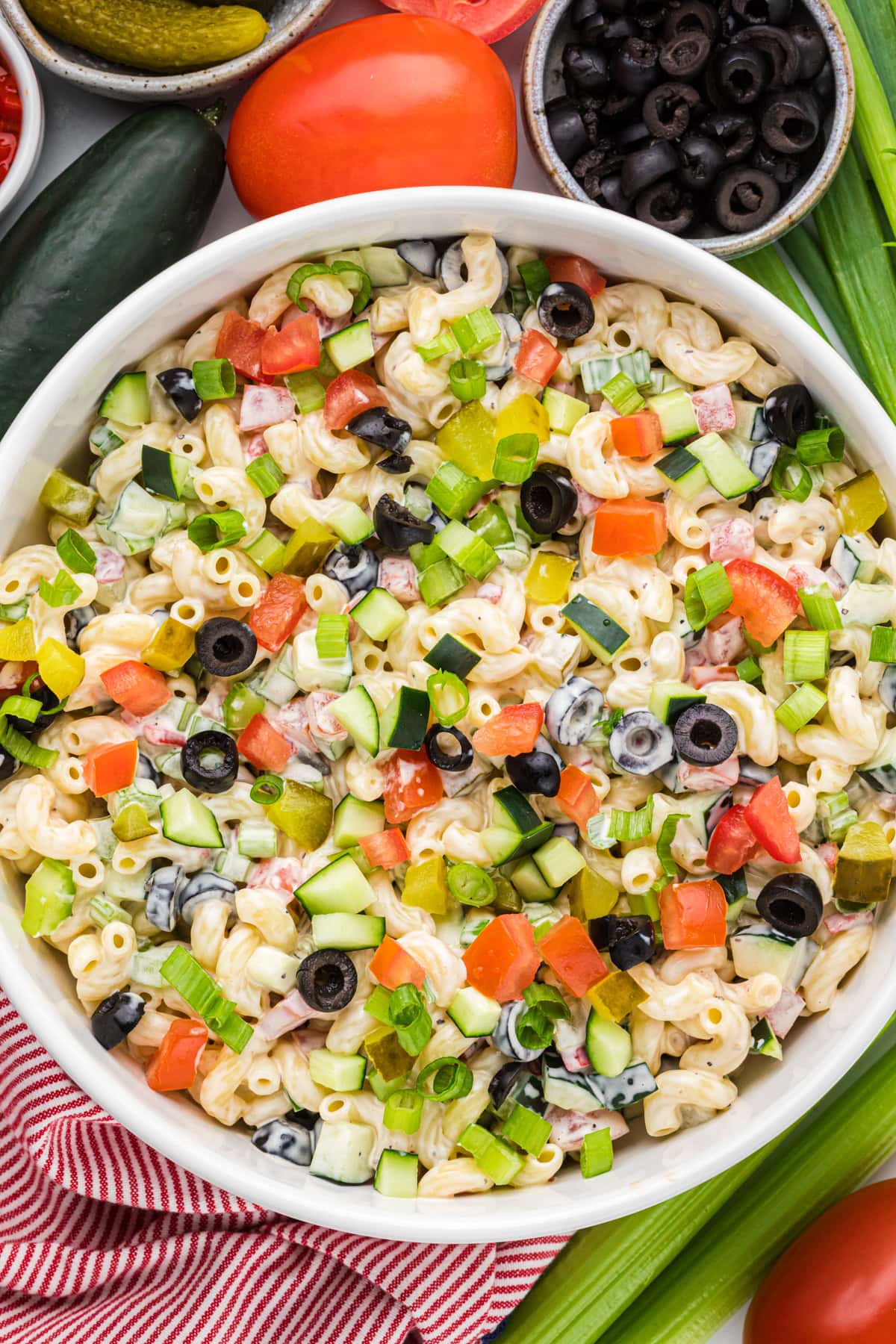 A bowl of macaroni salad mixed with diced cucumbers, tomatoes, green onions, pickles, black olives, and a creamy dressing—an ideal choice for side dishes at your graduation party. Surrounding ingredients are visible at the edges.