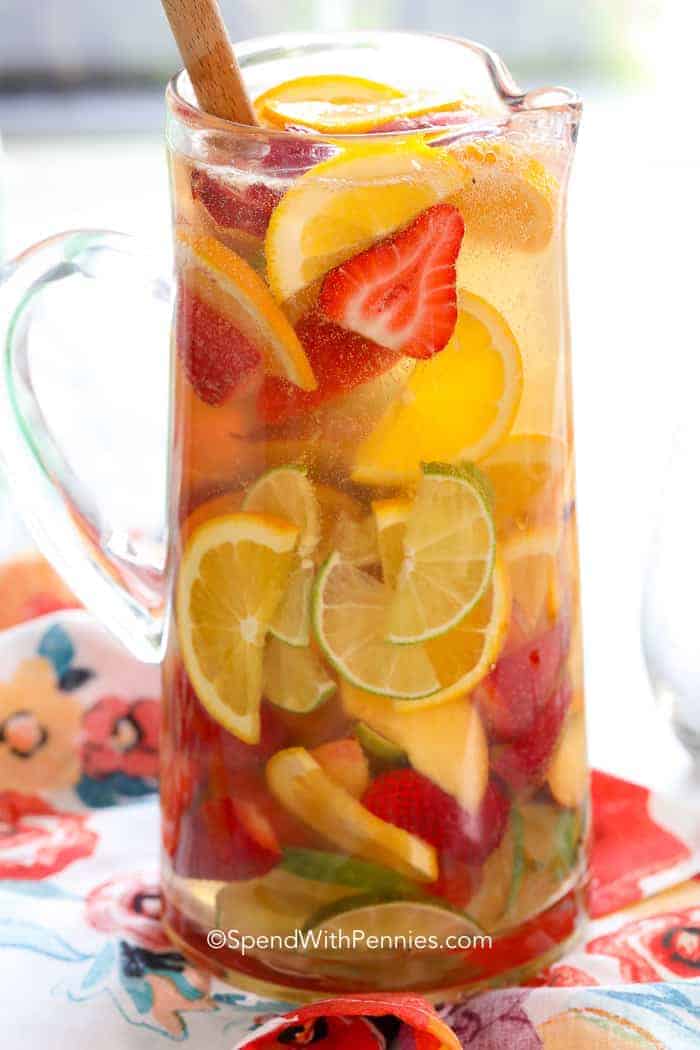 A glass pitcher filled with summer sangria, containing sliced lemons, limes, oranges, and strawberries, with a wooden spoon inside.