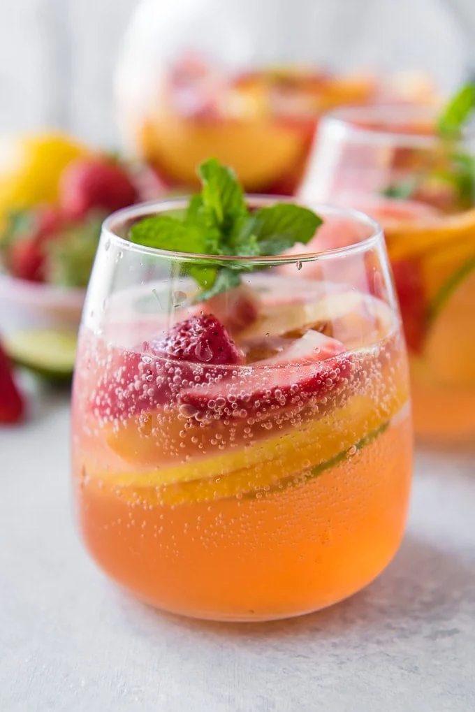 A glass of summer sangria-inspired sparkling fruit drink with ice, sliced strawberries, lemon, and cucumber, garnished with a sprig of mint.