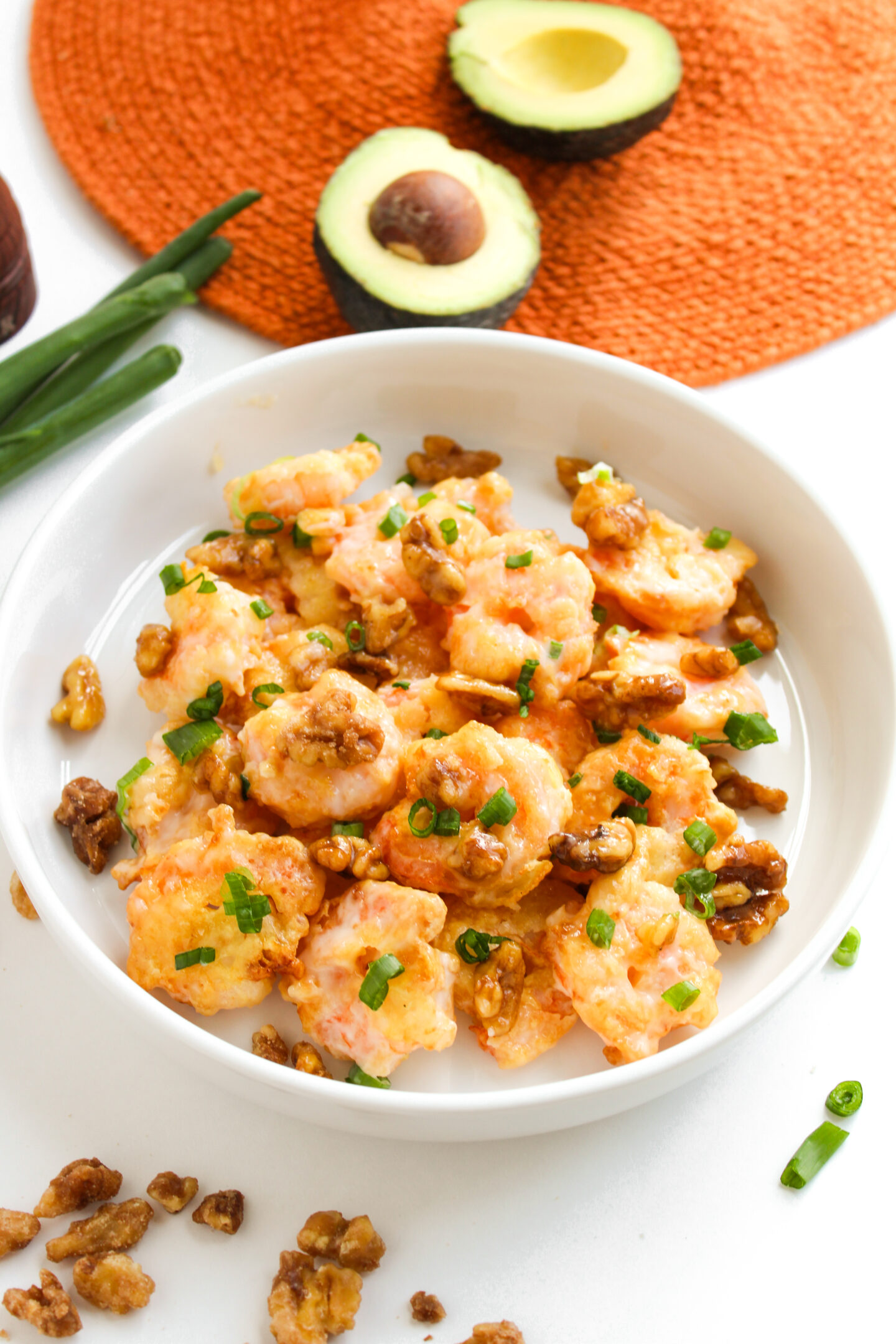 A white bowl filled with creamy shrimp, perfect for a summer dinner, topped with chopped green onions and walnuts, with avocado halves and fresh green onions in the background.