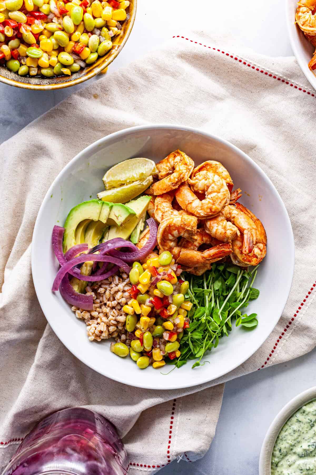 A white bowl containing shrimp, avocado slices, lime wedges, red onion, corn salad, microgreens, and farro makes a perfect high protein summer meal on a beige cloth.