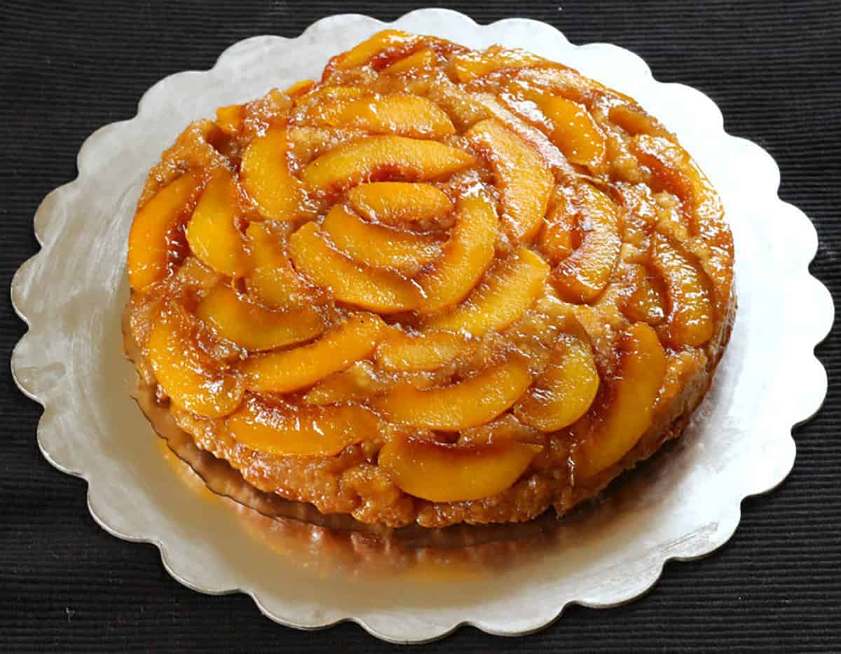 A peach dessert featuring an upside-down cake with caramelized peach slices arranged in a circular pattern on a scalloped metal platter.