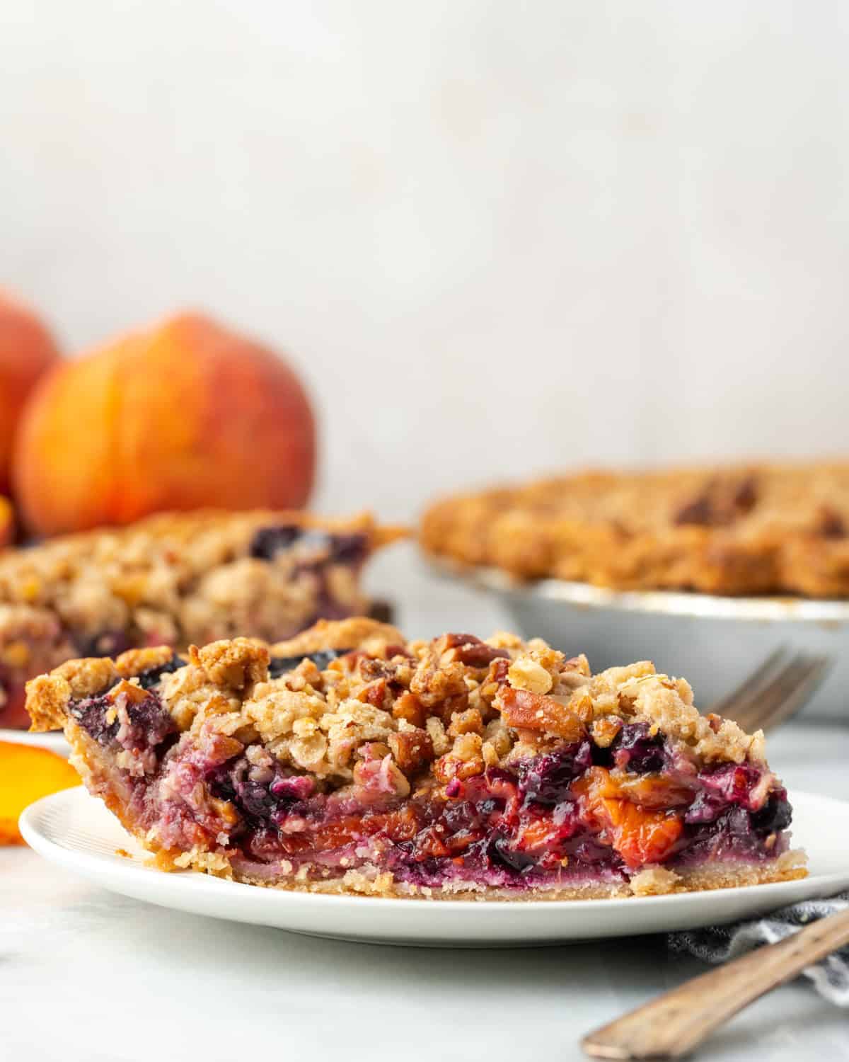 A slice of fruit pie with crumble topping on a white plate, featuring whole peaches and the rest of the peach dessert in the background.