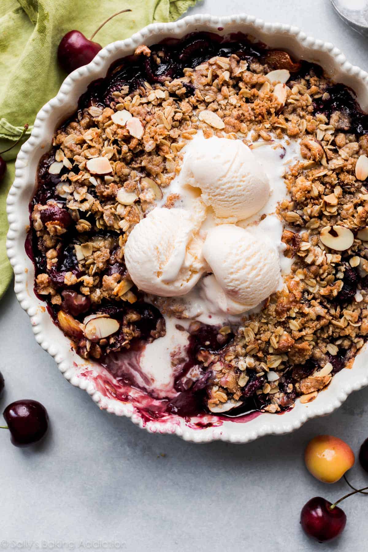 A classic summer dessert, this baked cherry crisp is topped with three scoops of vanilla ice cream, served in a white pie dish with fresh cherries and a green napkin nearby.