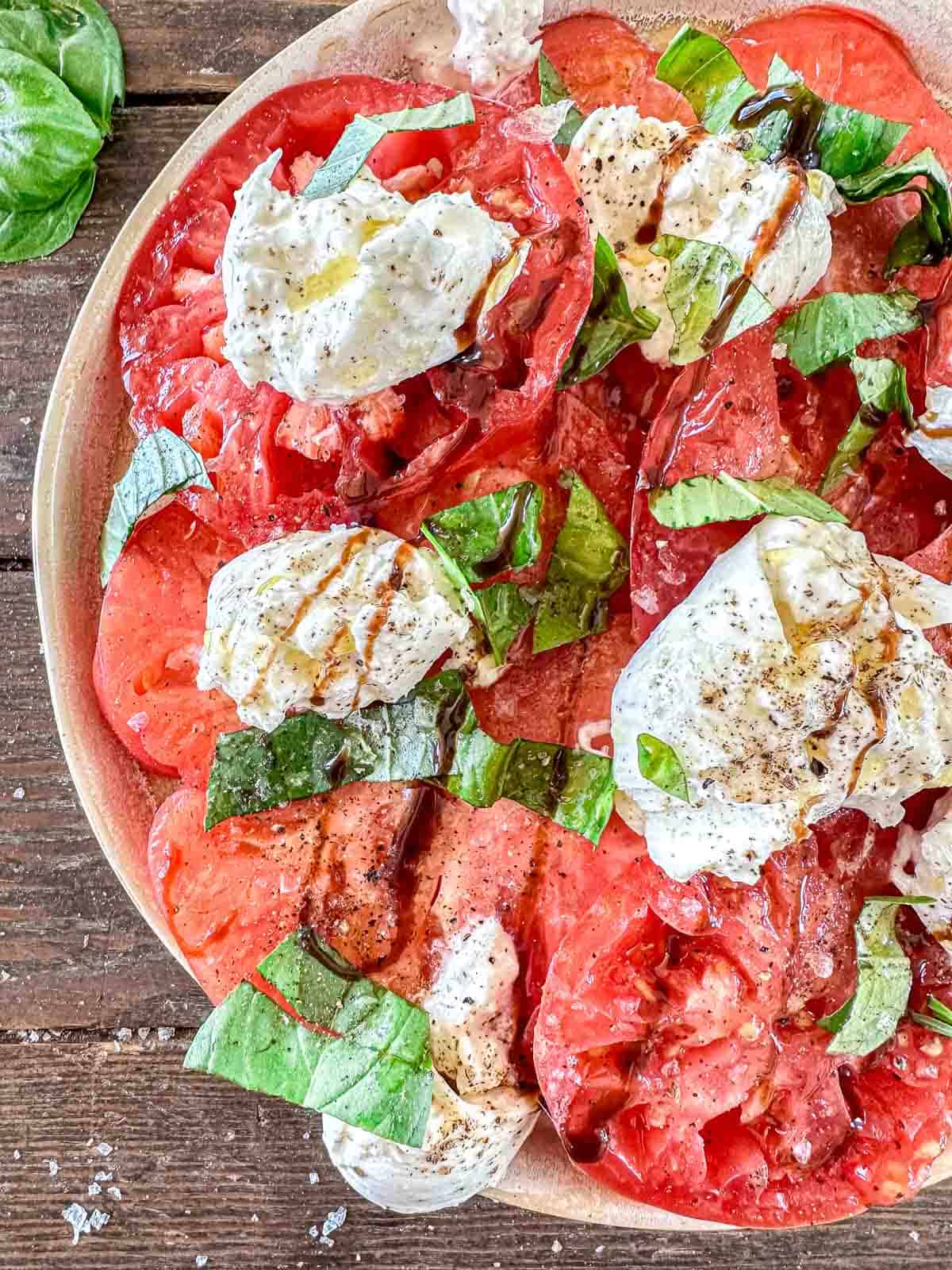 A plate of sliced tomatoes topped with burrata cheese, fresh basil leaves, cracked black pepper, and a drizzle of balsamic glaze makes this dish a classic from Italian Summer Recipes, perfect for enjoying on a wooden surface.