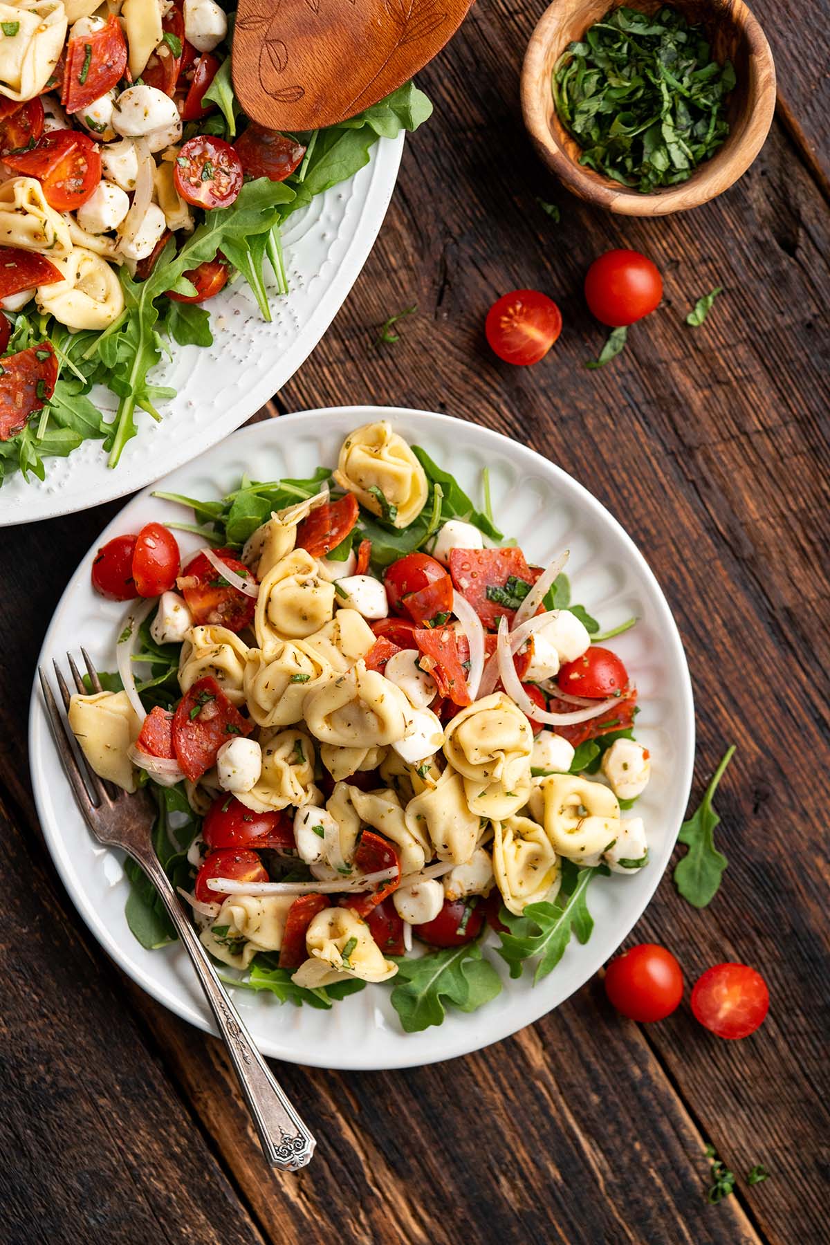 A plate of tortellini salad with cherry tomatoes, mozzarella balls, onion, and arugula on a wooden table—an ideal side dish for a graduation party—with a fork and a small bowl of chopped herbs nearby.