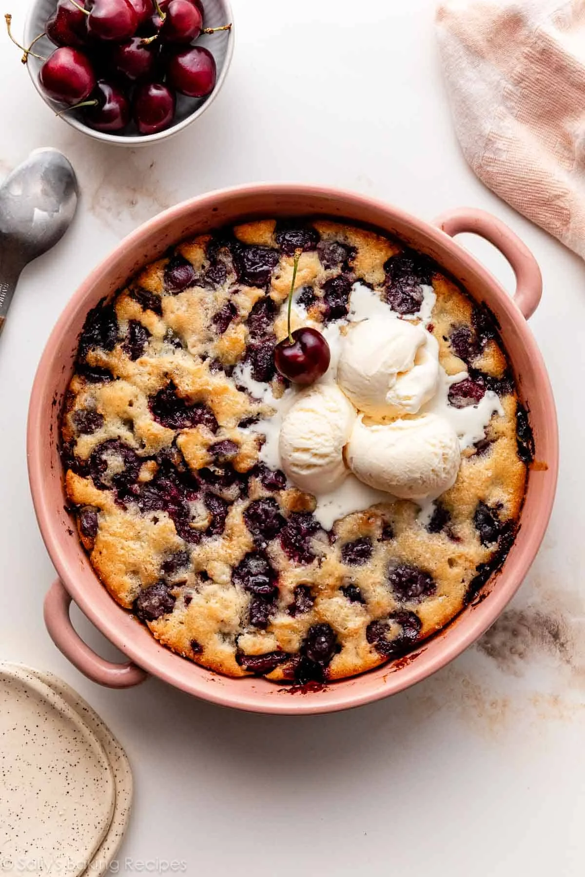 A cherry cobbler in a pink baking dish topped with three scoops of vanilla ice cream and a cherry, with a bowl of cherries and a spoon nearby—an irresistible summer dessert.
