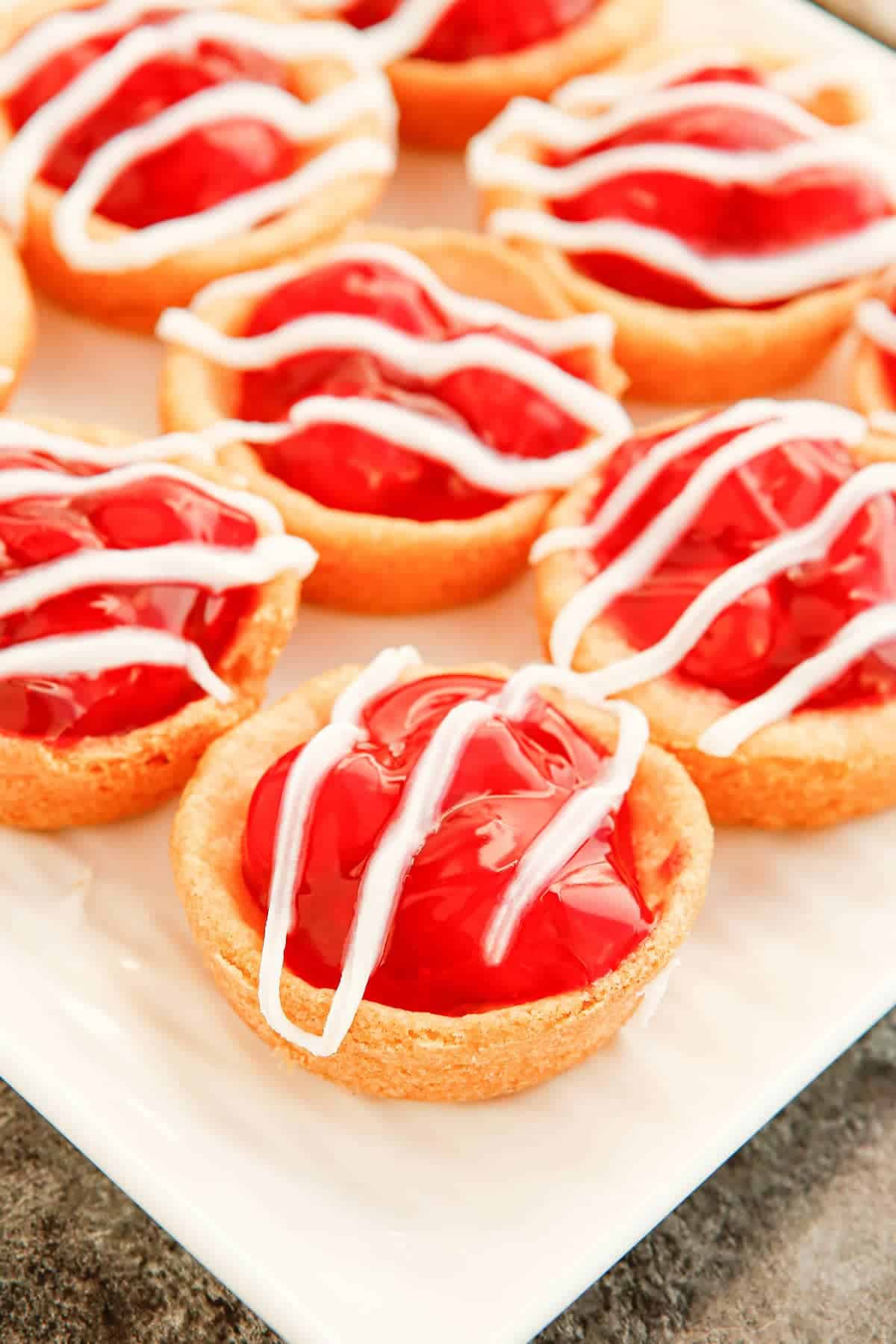 Mini cherry pies with a golden crust, filled with bright red cherry filling and drizzled with white icing, arranged on a white rectangular plate—perfect as a summer dessert.
