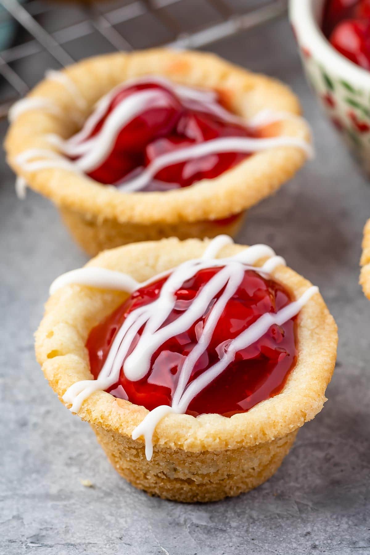 Two small summer dessert tarts filled with cherry pie filling and topped with a white icing drizzle, sitting on a gray surface.