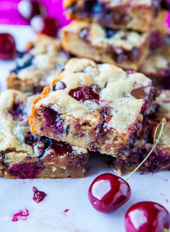 Cherry chocolate chip cookie bars stacked on parchment paper, with fresh cherries scattered around—an irresistible summer dessert.