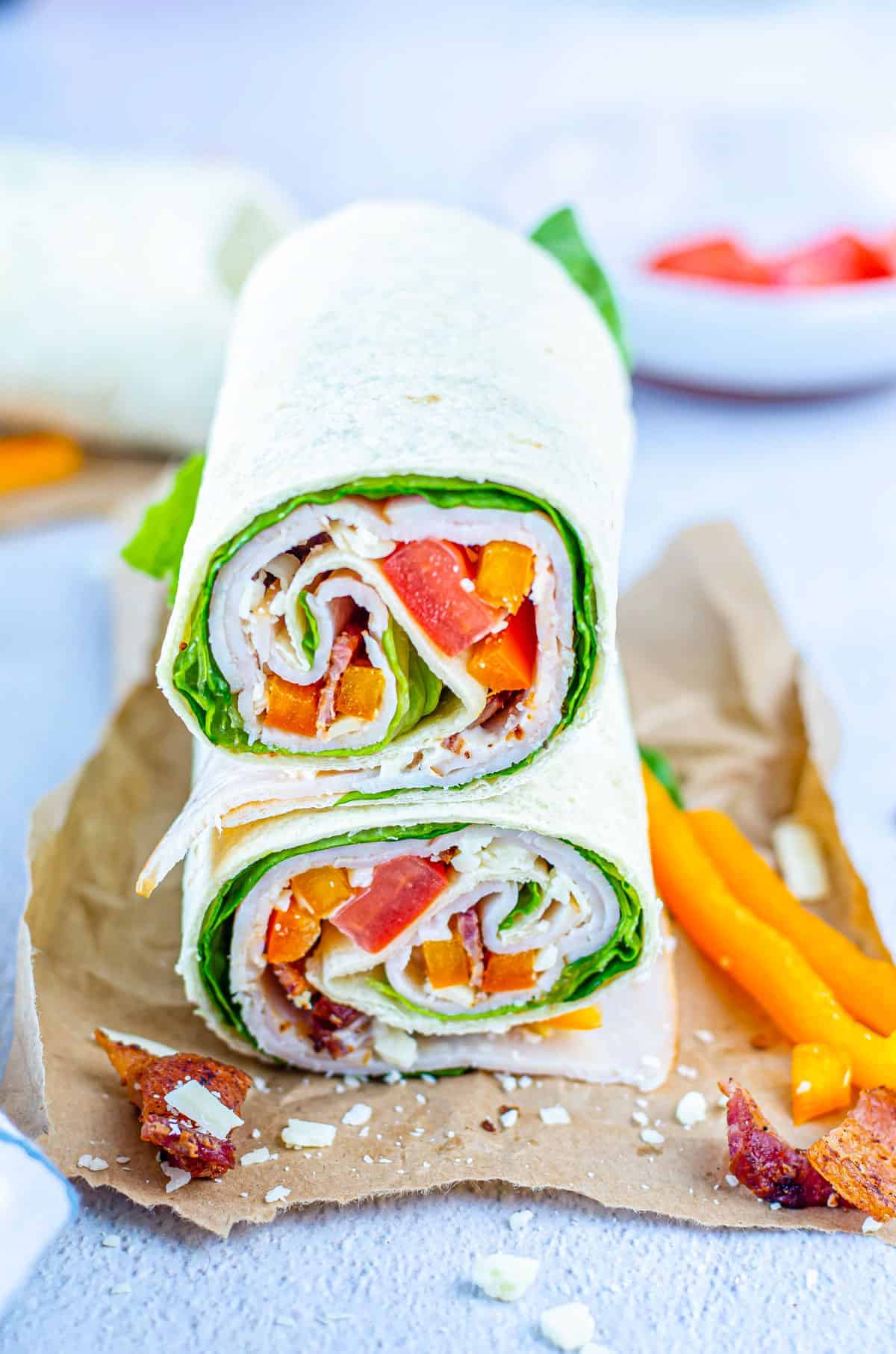 Two tortilla wraps filled with lettuce, turkey slices, diced tomatoes, and yellow bell peppers—stacked on brown paper with extra peppers and crumbs—make perfect beach picnic food for anyone seeking tasty lunch ideas without sandwiches.