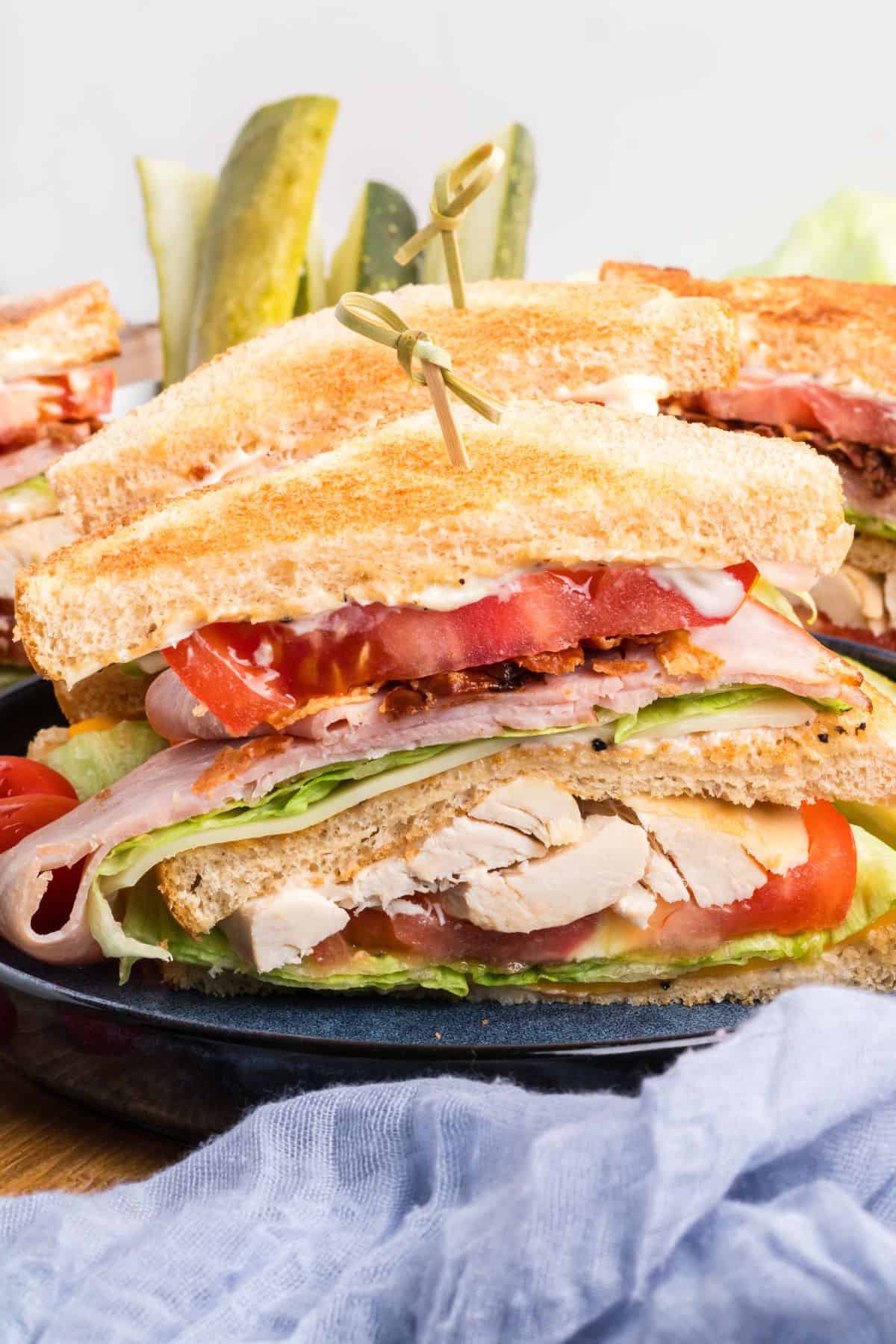 A close-up of a stacked club sandwich with turkey, bacon, lettuce, tomato, and mayonnaise on toasted bread, secured with toothpicks and served with pickle spears—perfect for summer snacks or quick kids lunch.