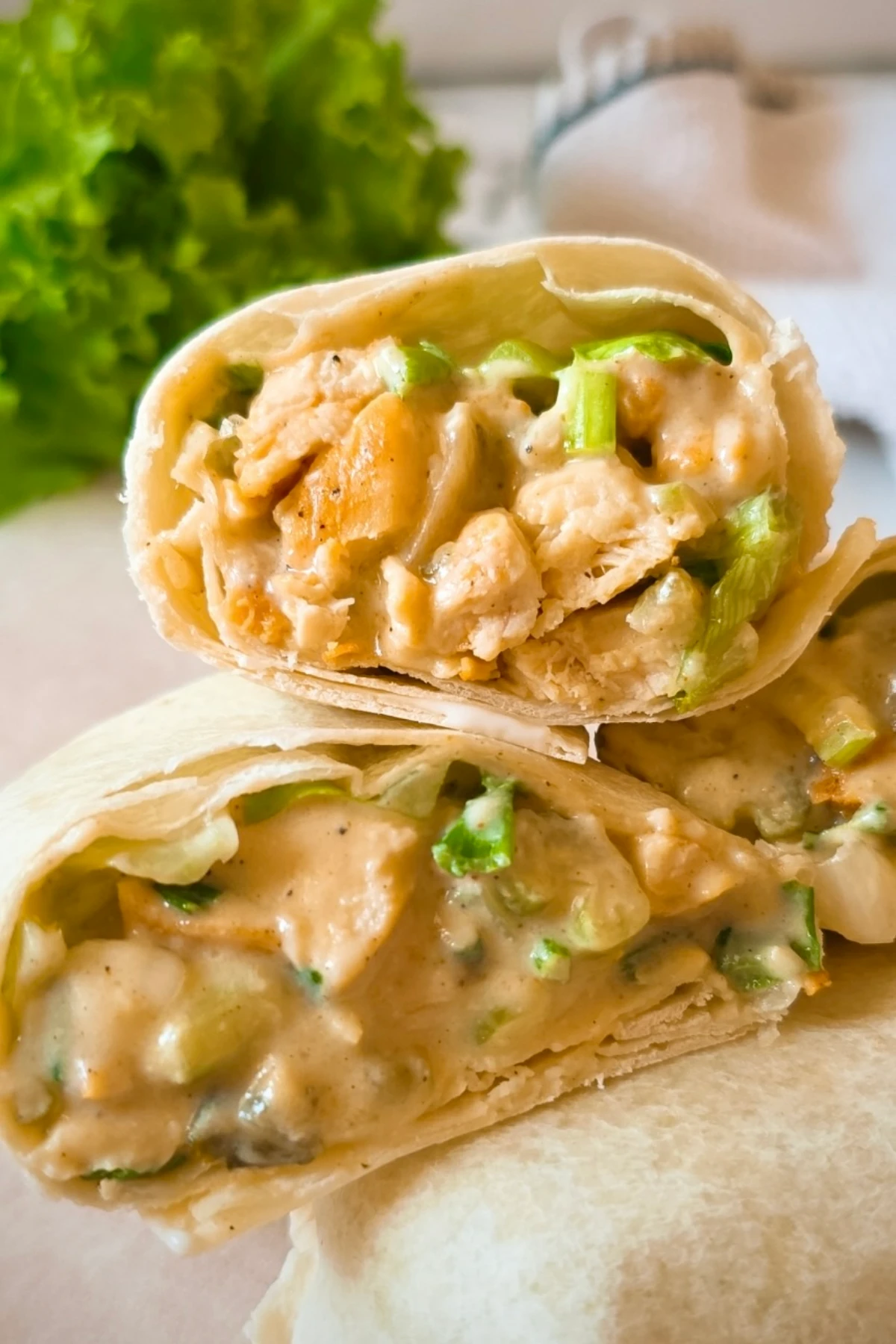 A close-up of a wrap cut in half, showing a creamy chicken and vegetable filling with visible green onions, on a light surface with lettuce in the background—perfect for cold beach lunch ideas.