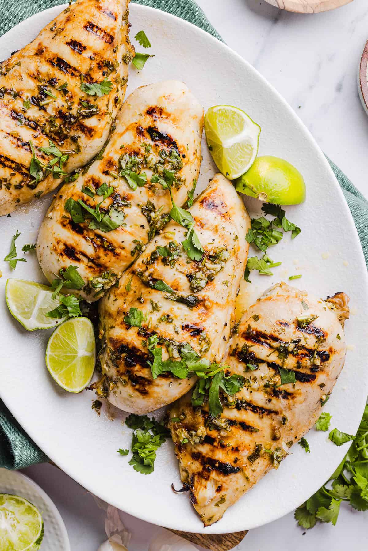 Four grilled chicken breasts garnished with chopped cilantro are served on a white plate with lime wedges, making this dish a perfect choice for Healthy Grilled Dinners.