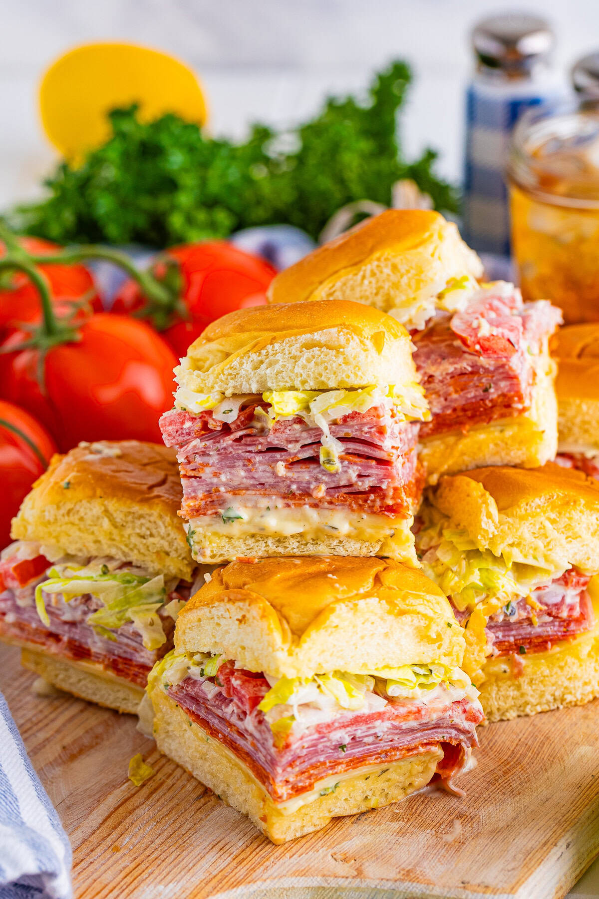 A stack of deli meat and cheese sliders with lettuce on soft buns, arranged on a wooden board with tomatoes and parsley in the background—perfect for pool lunch ideas or easy kids lunch.