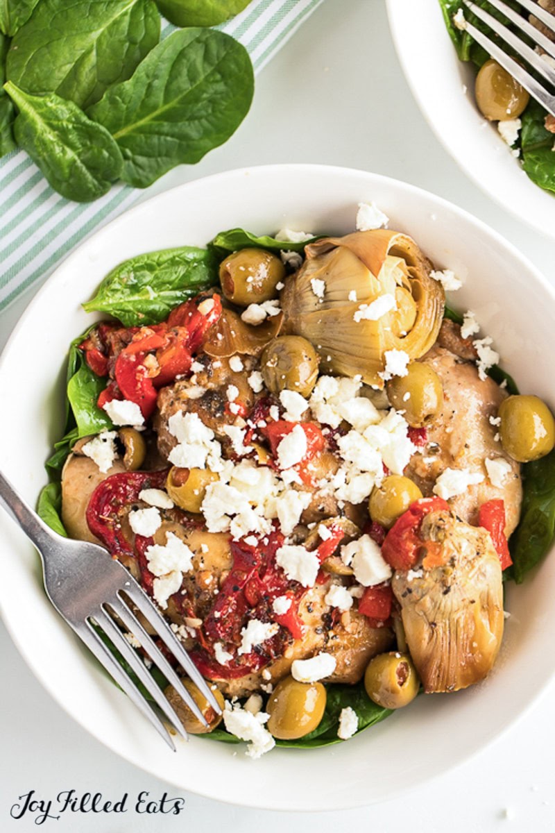 A white bowl filled with spinach, artichoke hearts, green olives, sun-dried tomatoes, grilled chicken, and crumbled feta cheese—a fresh twist perfect for summer recipes, with a fork resting on the side.