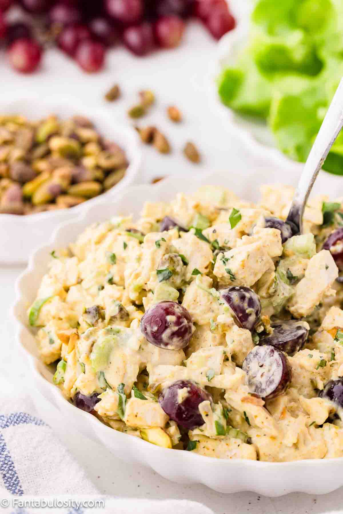A bowl of Chicken Salad with grapes, chopped herbs, and pistachios, perfect for Summer Recipes. A spoon rests in the bowl, with grapes, pistachios, and lettuce visible in the background.