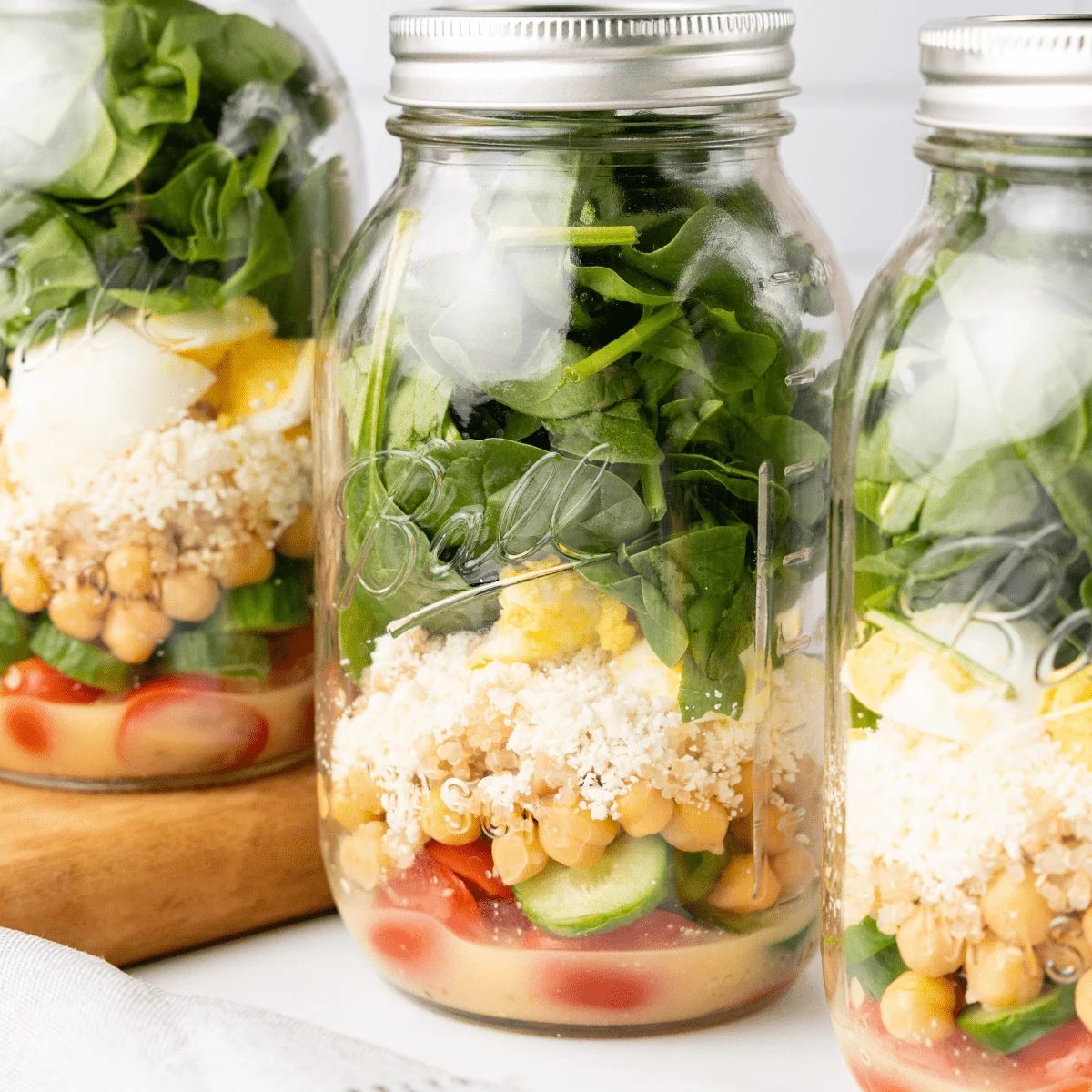 Three glass jars are filled with layered salad ingredients—spinach, eggs, feta cheese, chickpeas, cucumbers, and tomatoes—with dressing at the bottom. Perfect for boat food or cooler meals on a relaxing boat day.