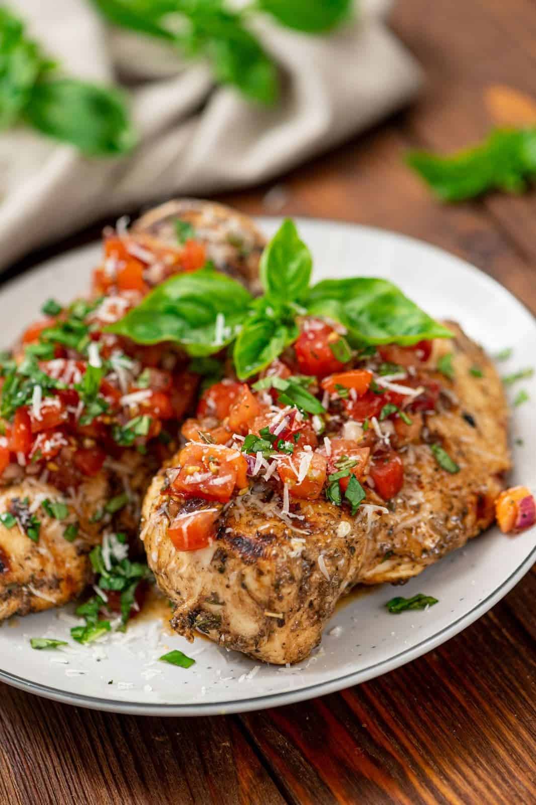 Grilled chicken breast topped with chopped tomatoes, basil, and grated cheese on a white plate, garnished with fresh herbs—perfect for those seeking Italian Summer Recipes.