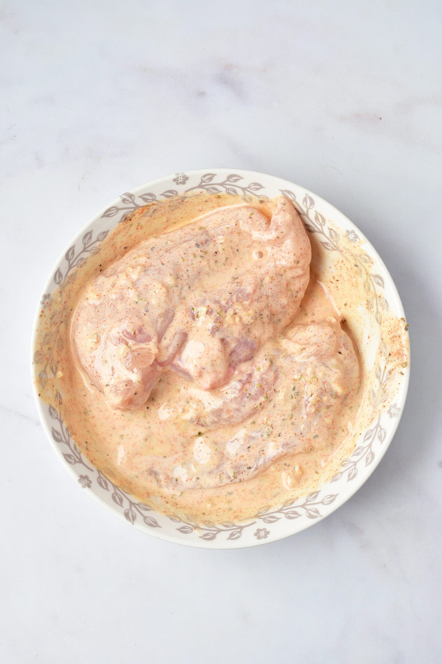 Two raw chicken breasts are coated in a creamy, seasoned marinade inside a white bowl with a decorative rim, ready to become flavorful Greek Chicken bowls, all placed on a light surface.
