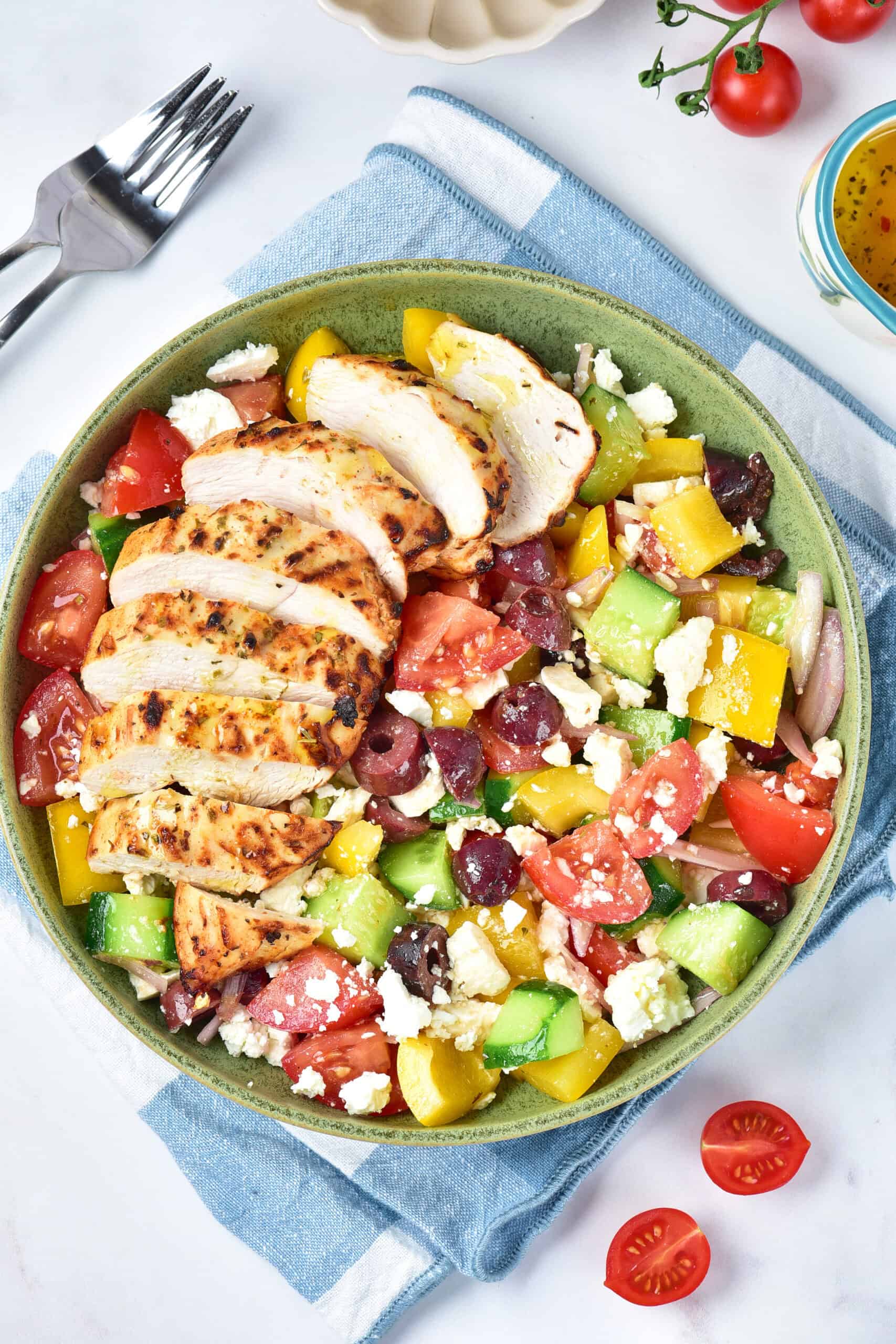 A green bowl with sliced grilled chicken breast served over a Greek salad with tomatoes, cucumbers, bell peppers, olives, red onions, and feta cheese. A fork and napkin are beside the bowl.