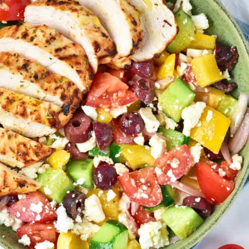 A bowl of Greek salad with chopped tomatoes, cucumbers, bell peppers, olives, feta cheese, red onion, and sliced Air Fryer Greek Chicken breast on top.