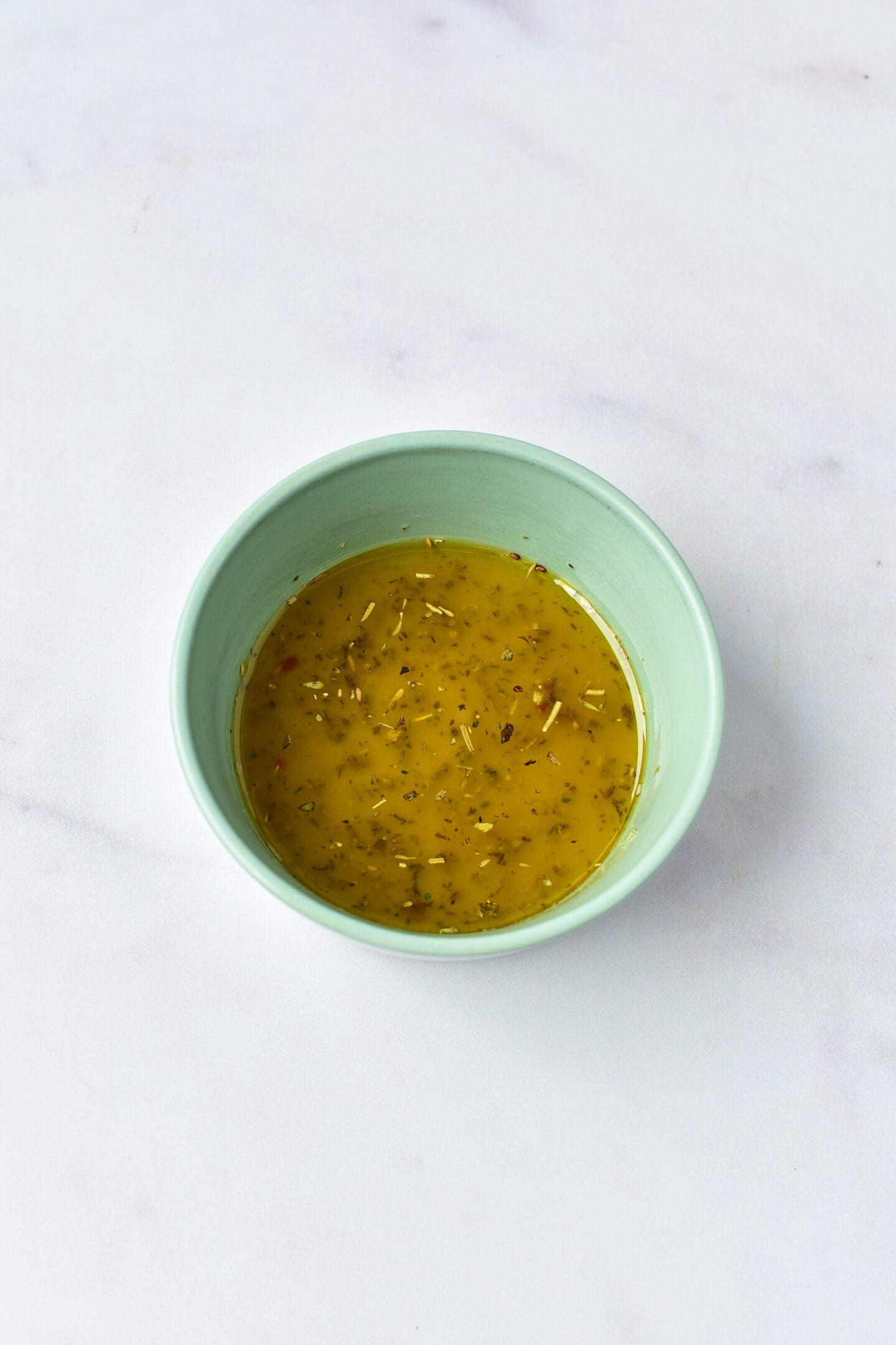A light green bowl filled with a yellow, herb-flecked Greek Chicken broth sits on a white marble surface.