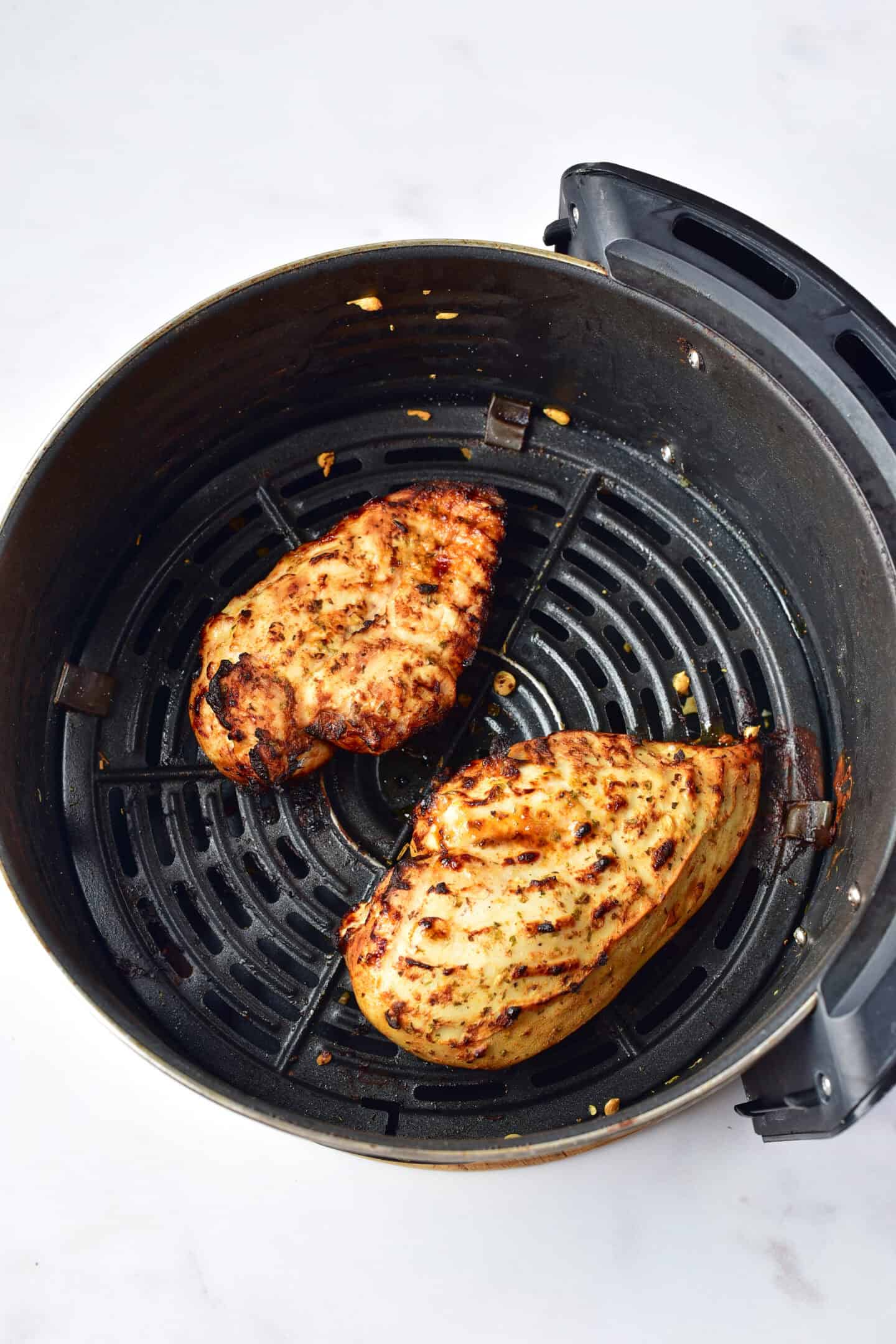 Two Greek Chicken breasts in an air fryer basket, cooked to perfection and lightly charred, rest on a white surface.