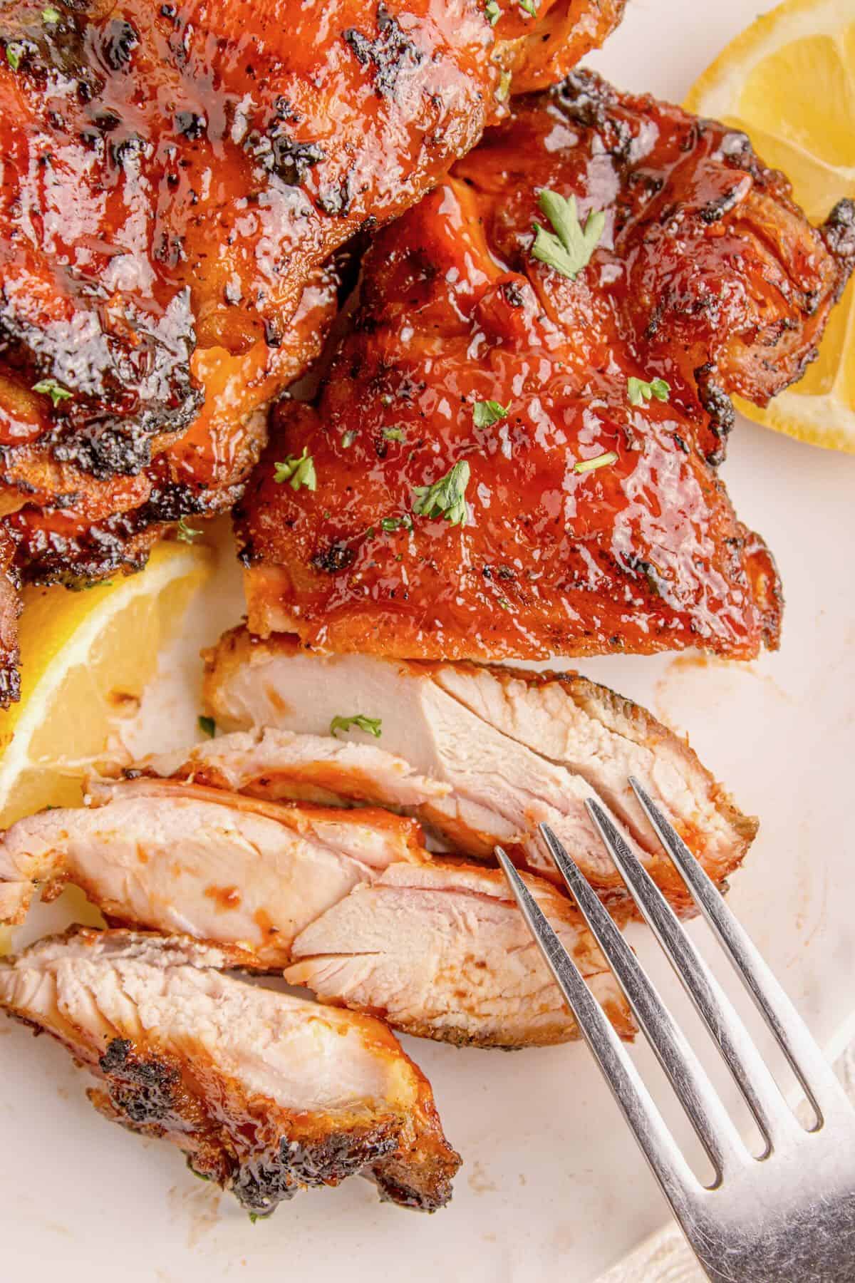 Close-up of succulent summer grilled chicken thighs glazed and garnished with fresh herbs, served with lemon wedges; a fork rests beside perfectly sliced pieces of chicken.