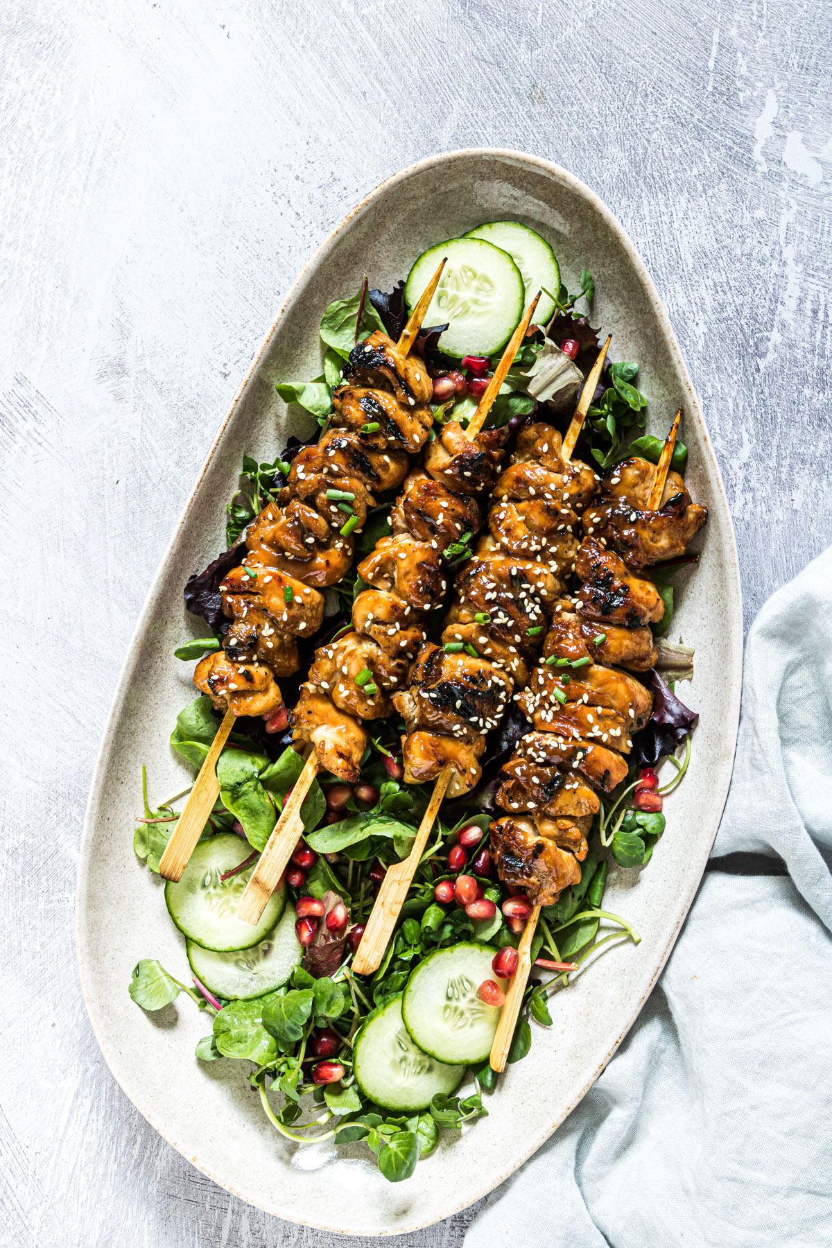 Three grilled chicken skewers with sesame seeds on a bed of mixed greens, cucumber slices, and pomegranate seeds capture the flavors of summer, served beautifully on an oval plate.