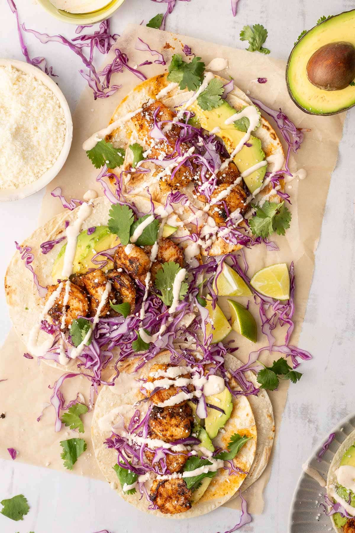 Three shrimp tacos on tortillas topped with shredded purple cabbage, avocado slices, cilantro, cheese, and a drizzle of sauce create a healthy grilled dinner. Lime wedges and half an avocado are nearby.