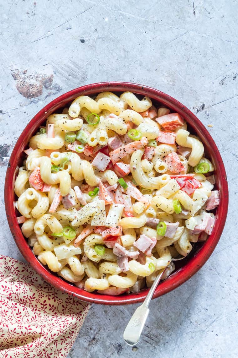 A red bowl filled with creamy pasta salad containing ham, bell peppers, and green onions—an irresistible side dish perfect for any graduation party or festive gathering, with a fork resting in the bowl.