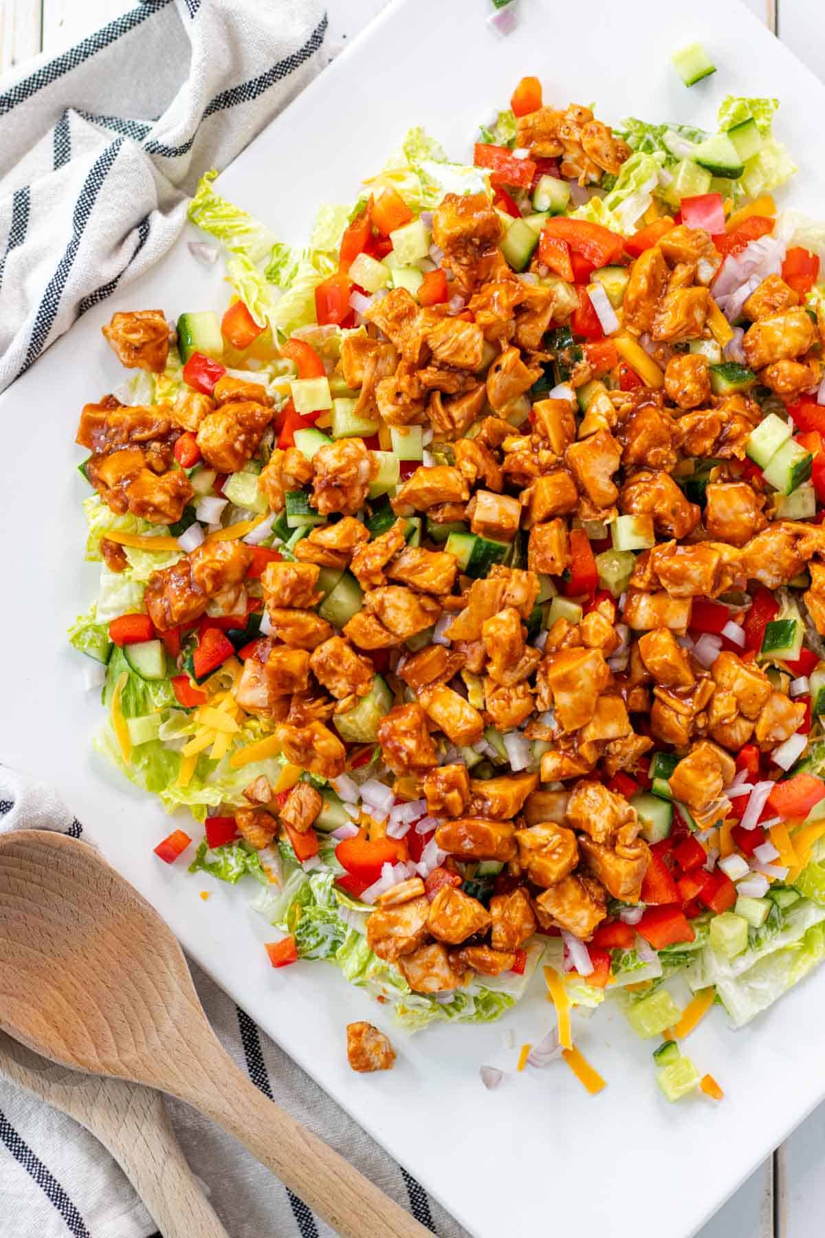 Chopped chicken salad with barbecue chicken, diced red onions, bell peppers, cucumbers, and lettuce on a white plate—perfect for summer recipes—served next to a striped towel and wooden spoons.