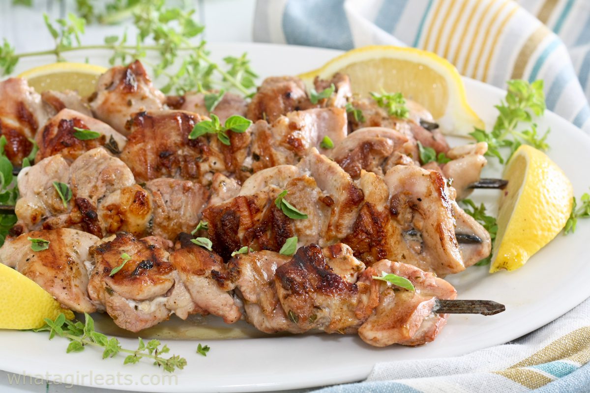 Grilled chicken skewers garnished with fresh herbs are served on a white plate with lemon wedges and leafy greens, perfect for summer grilling.