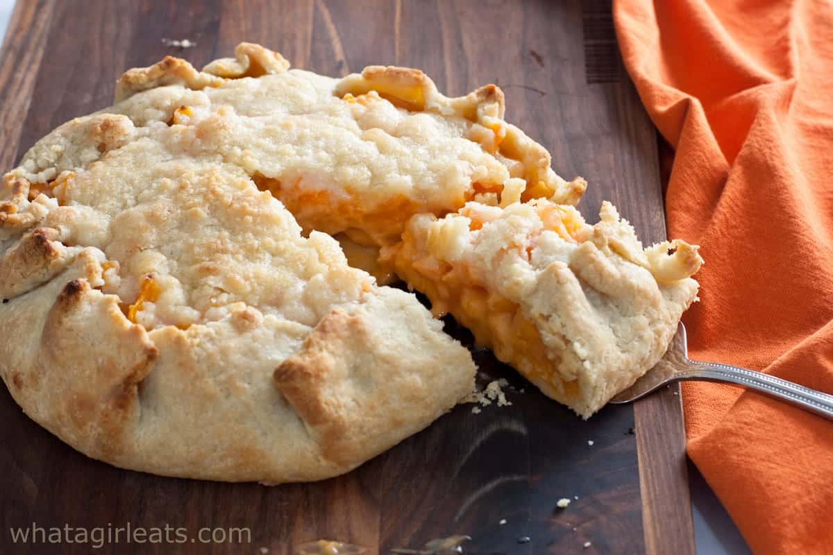 A rustic, open-faced savory pie with a flaky crust and creamy orange filling sits on a wooden board, resembling a peach dessert; one slice is being lifted out. An orange cloth is next to it.
