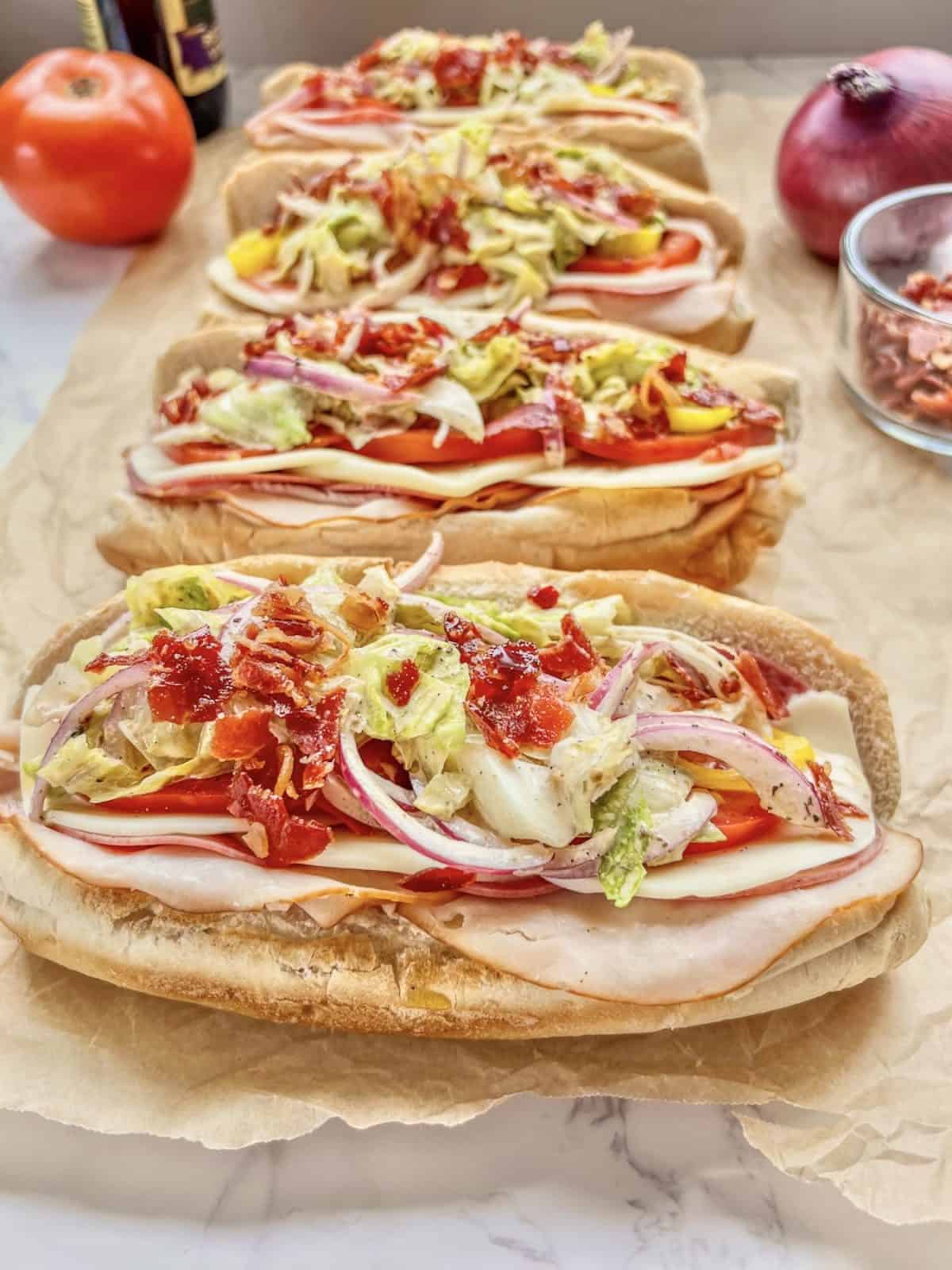 Four sub sandwiches on parchment paper topped with sliced turkey, cheese, tomatoes, lettuce, red onion, and bacon bits—a fresh idea for Italian Summer Recipes—with a tomato and red onion in the background.