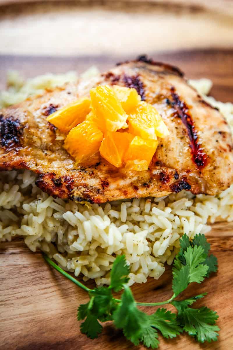 Grilled chicken breast, perfect for summer, topped with chopped orange pieces and served on a bed of seasoned rice, garnished with cilantro on a wooden plate.