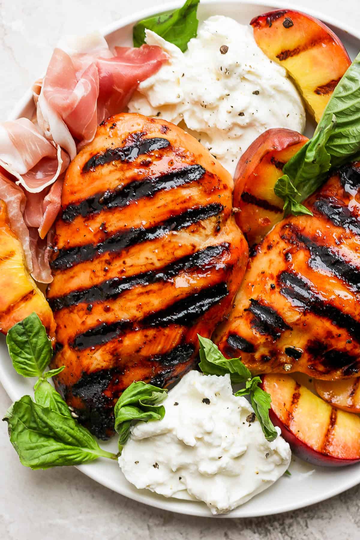 Grilled chicken breasts with char marks, served with burrata cheese, juicy summer peach slices, prosciutto, and fresh basil leaves on a white plate.