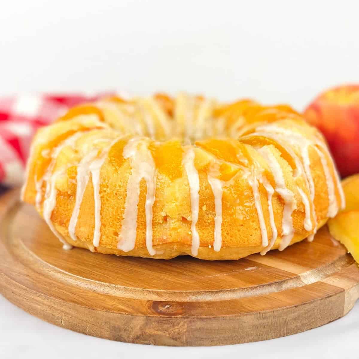A glazed peach dessert bundt cake with peach slices sits on a round wooden board, accompanied by a red checkered cloth and a fresh peach in the background.