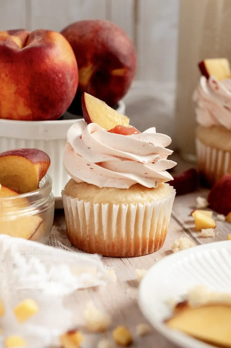 A vanilla cupcake topped with peach-flavored frosting and a peach slice, surrounded by fresh peaches and peach pieces on a wooden surface—a delightful peach dessert for any occasion.