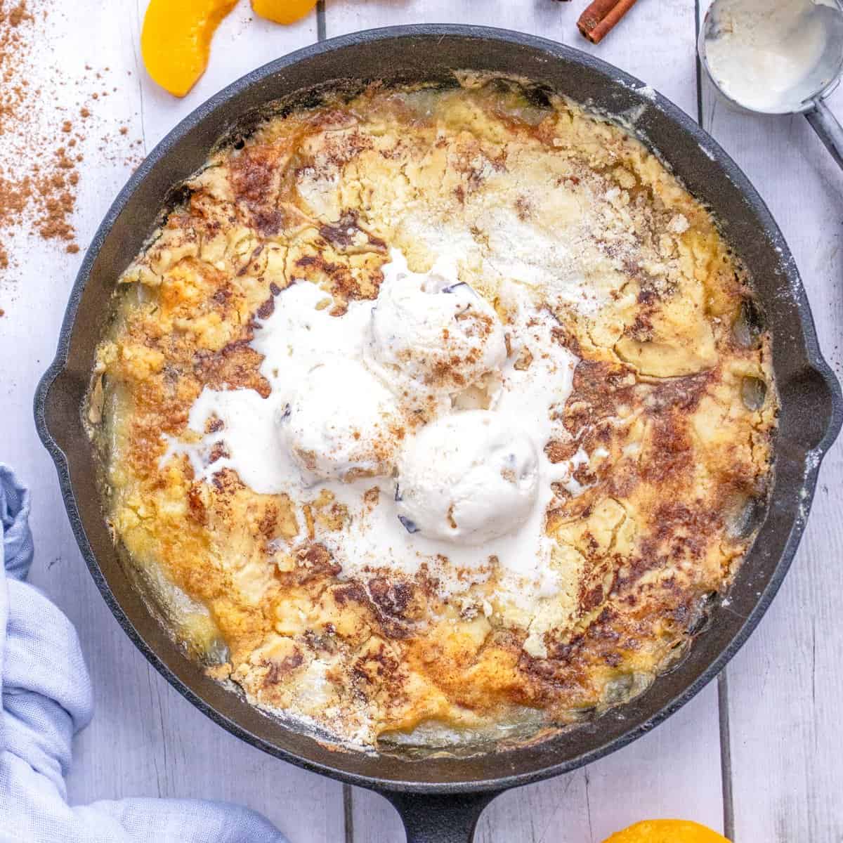 A cast iron skillet filled with peach dessert cobbler, topped with three scoops of melting vanilla ice cream, on a white wooden surface.